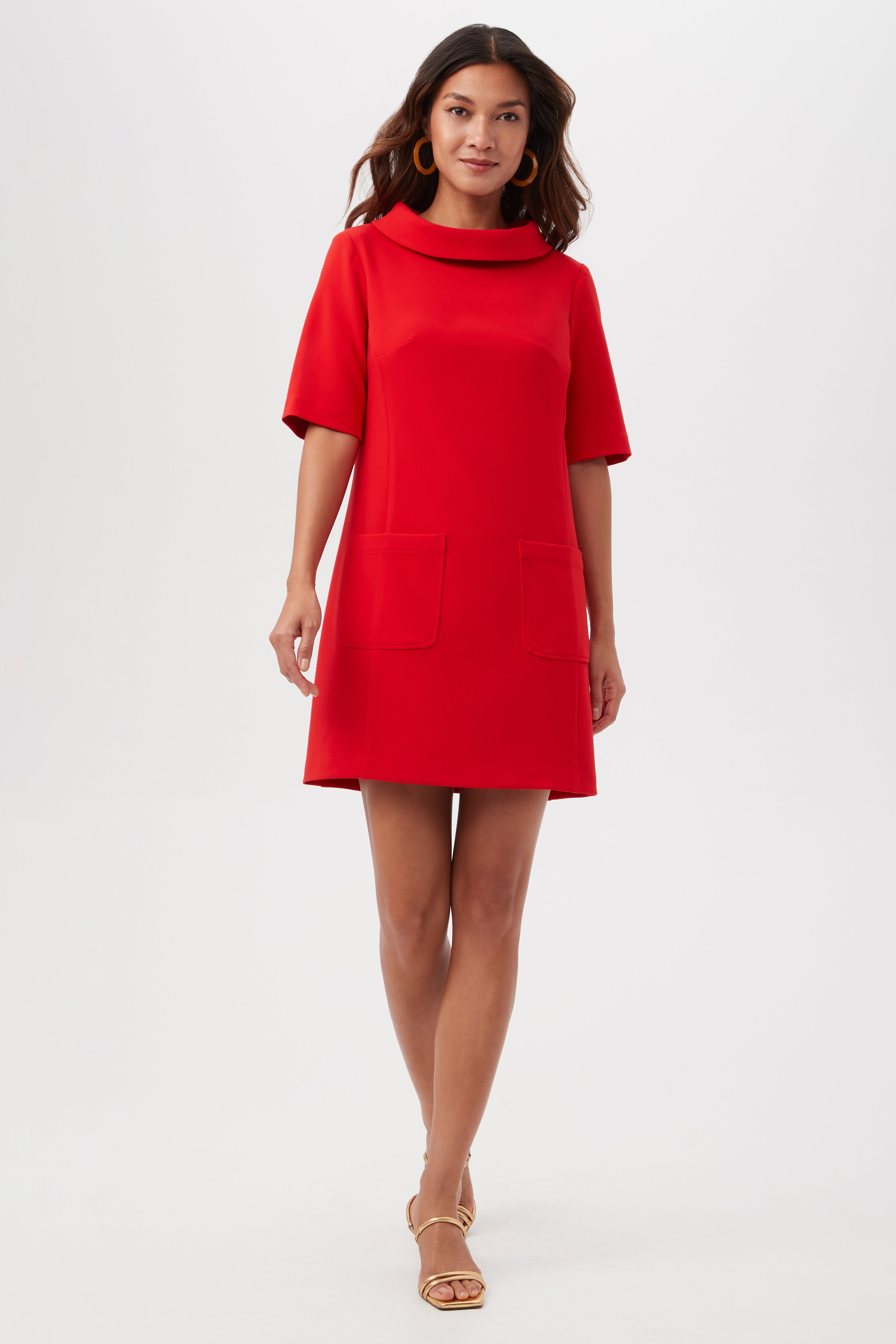 A woman models the Maleko Dress, a short-sleeved, bright red mini dress with a high neckline and front pockets, styled with gold open-toe sandals against a plain white background.
