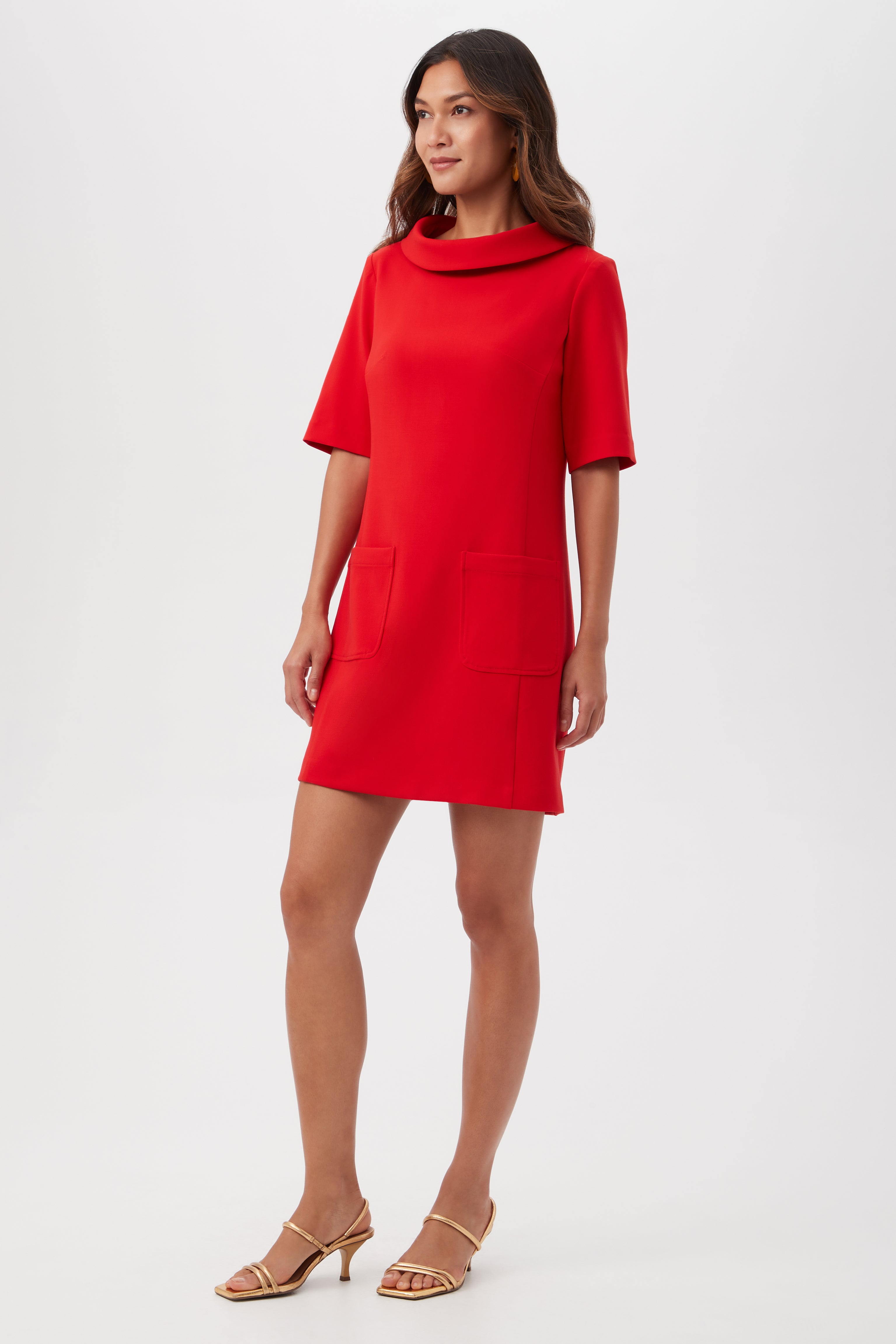 A woman stands in heels against a plain white background, wearing the Maleko Dress—a bright red, short-sleeve mini dress with a cowl neckline and two front pockets.