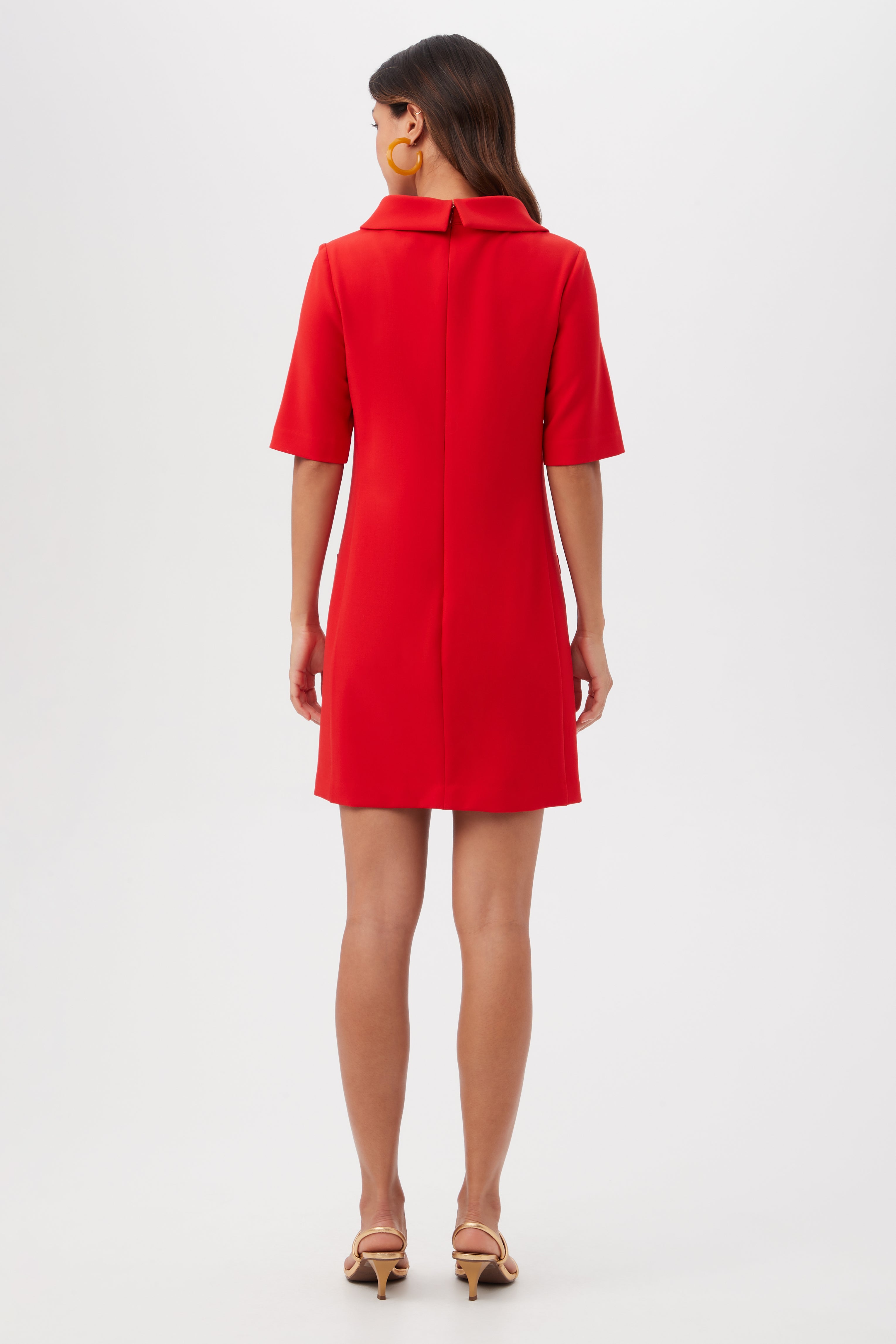 A woman with long brown hair and large round earrings faces away, modeling the Maleko Dress—a knee-length, short-sleeved red dress with a collar—and beige heeled sandals, against a plain white background.