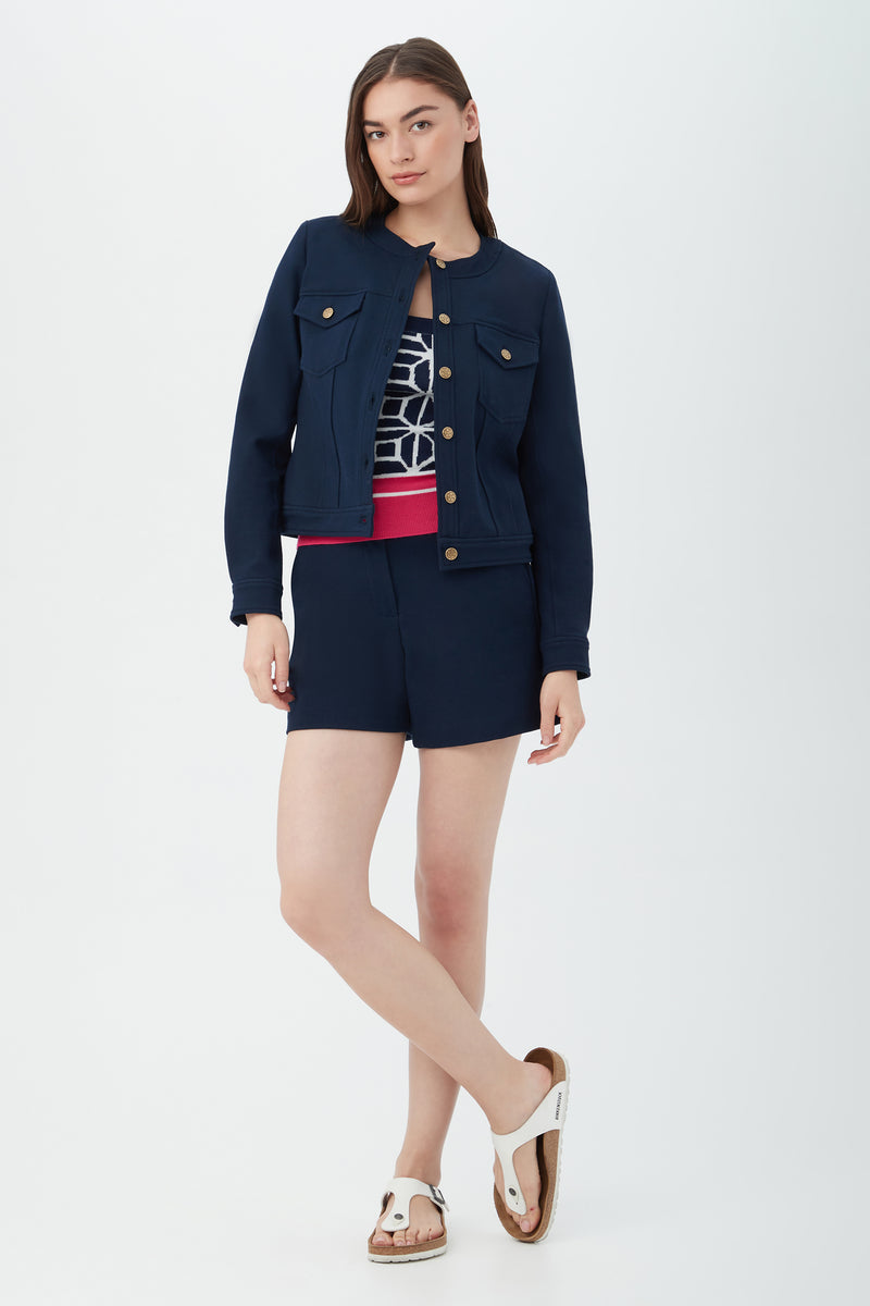 A woman with long brown hair stands in a relaxed pose against a white background, wearing Valera Shorts - Stretch Piqué in navy, paired with a matching jacket, patterned navy and white top with red waistband, and white sandals.