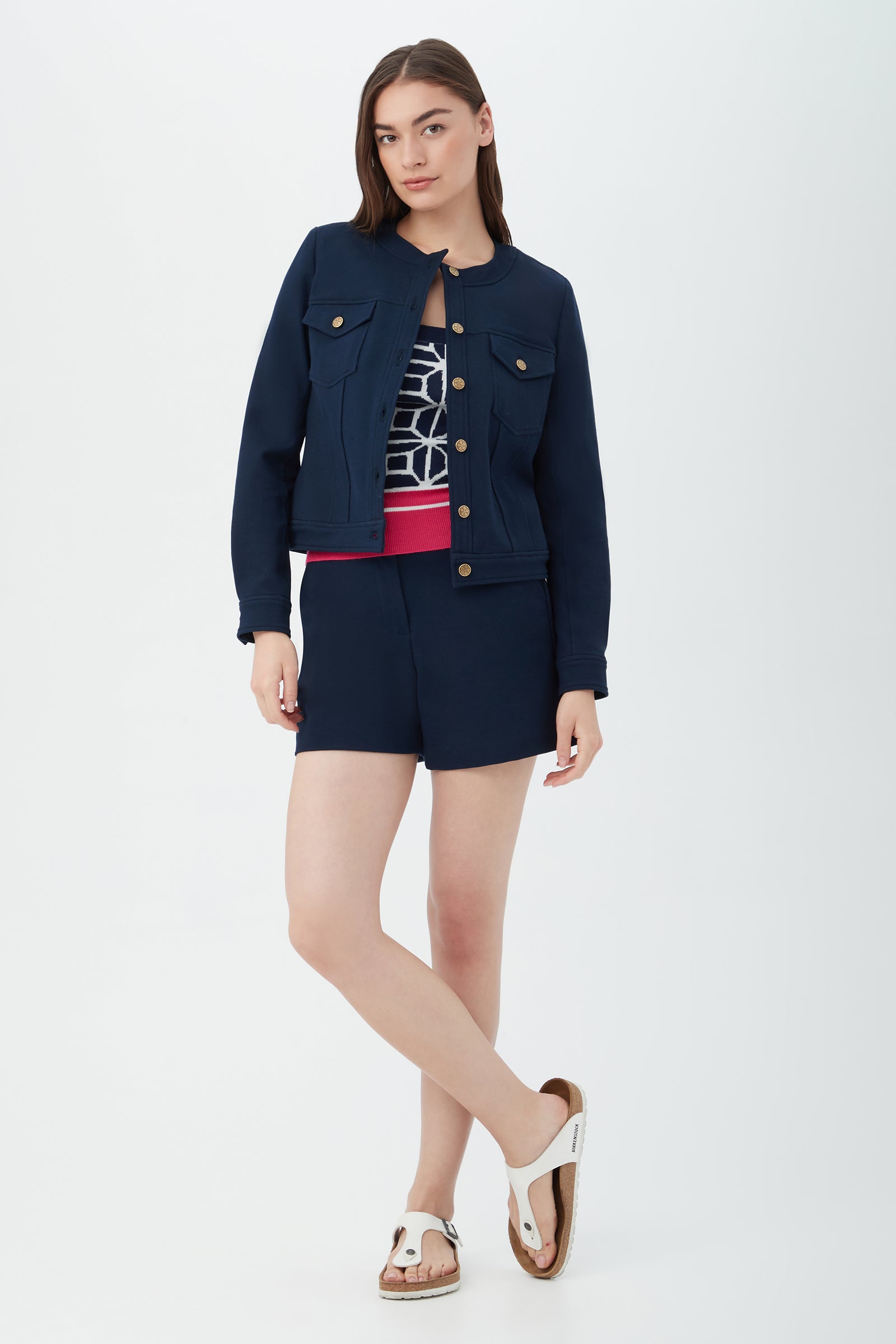 A woman with long straight hair poses casually against a white background, wearing the Gail Jacket in navy with matching shorts, a patterned top, and white sandals.
