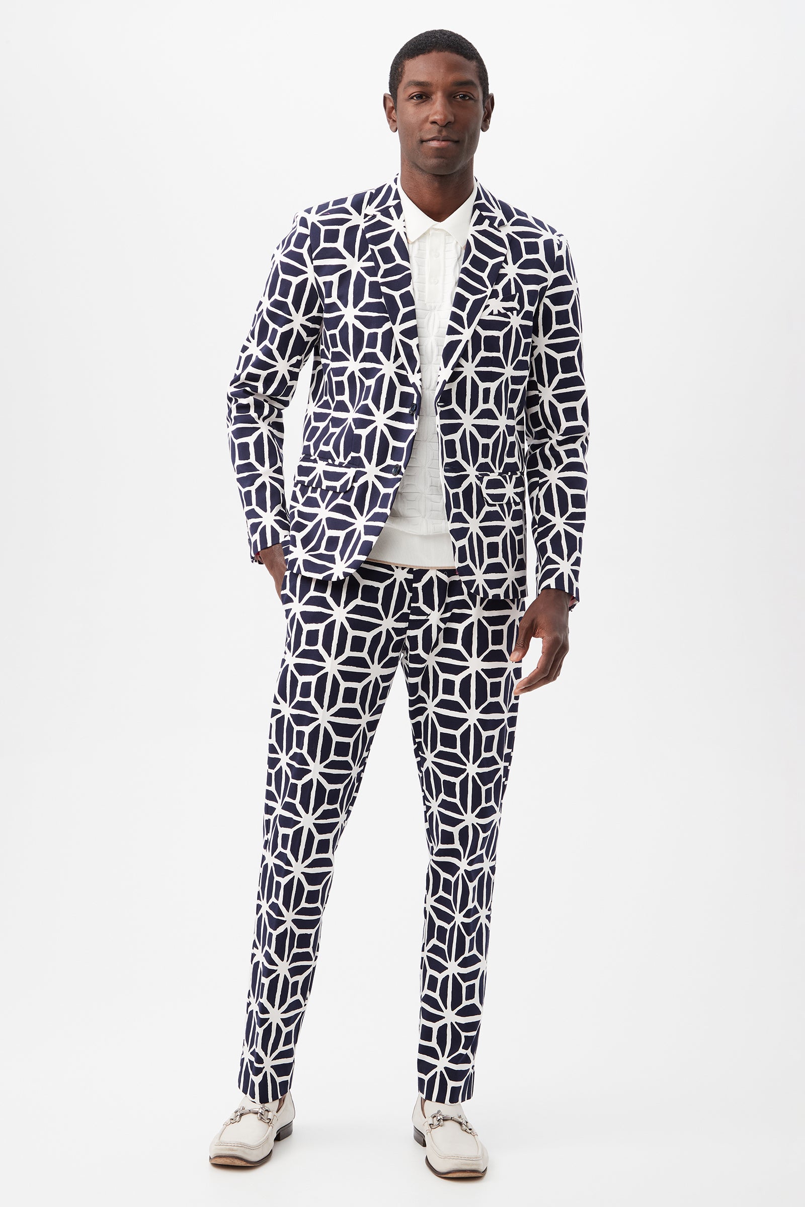 A man faces forward against a white background, wearing the Thurston Blazer - Durell Herringbone with a bold navy and white geometric pattern, paired with a white collared shirt and white shoes.