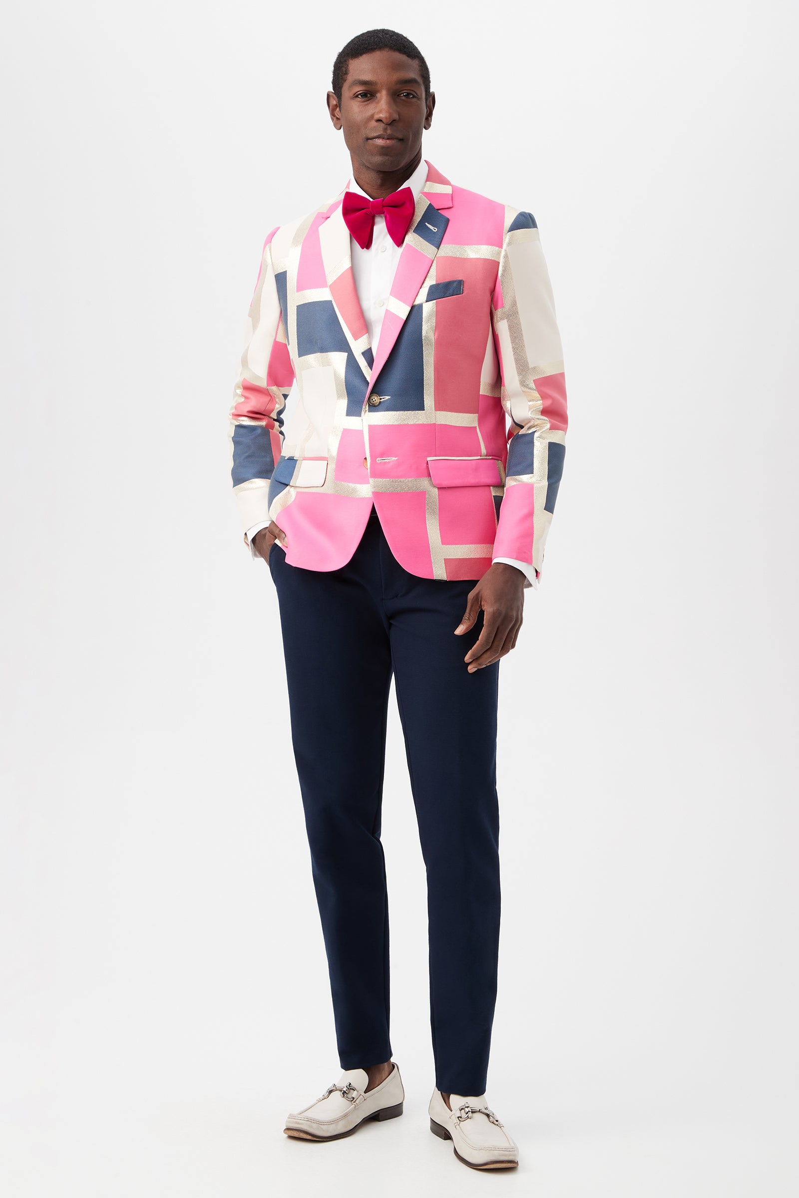 A man stands against a plain white background wearing the Thurston Blazer - Block Party Jacquard, featuring bold pink and blue geometric patterns, paired with a white shirt, red bow tie, navy dress pants, and white dress shoes.