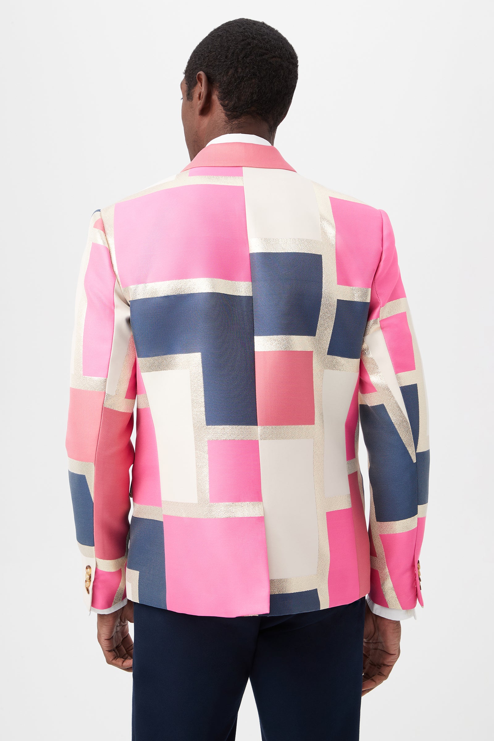 A person with short hair faces away, modeling the Thurston Blazer - Block Party Jacquard, featuring a vibrant geometric pattern in pink, cream, navy blue, and gold with large intersecting rectangles against a plain background.