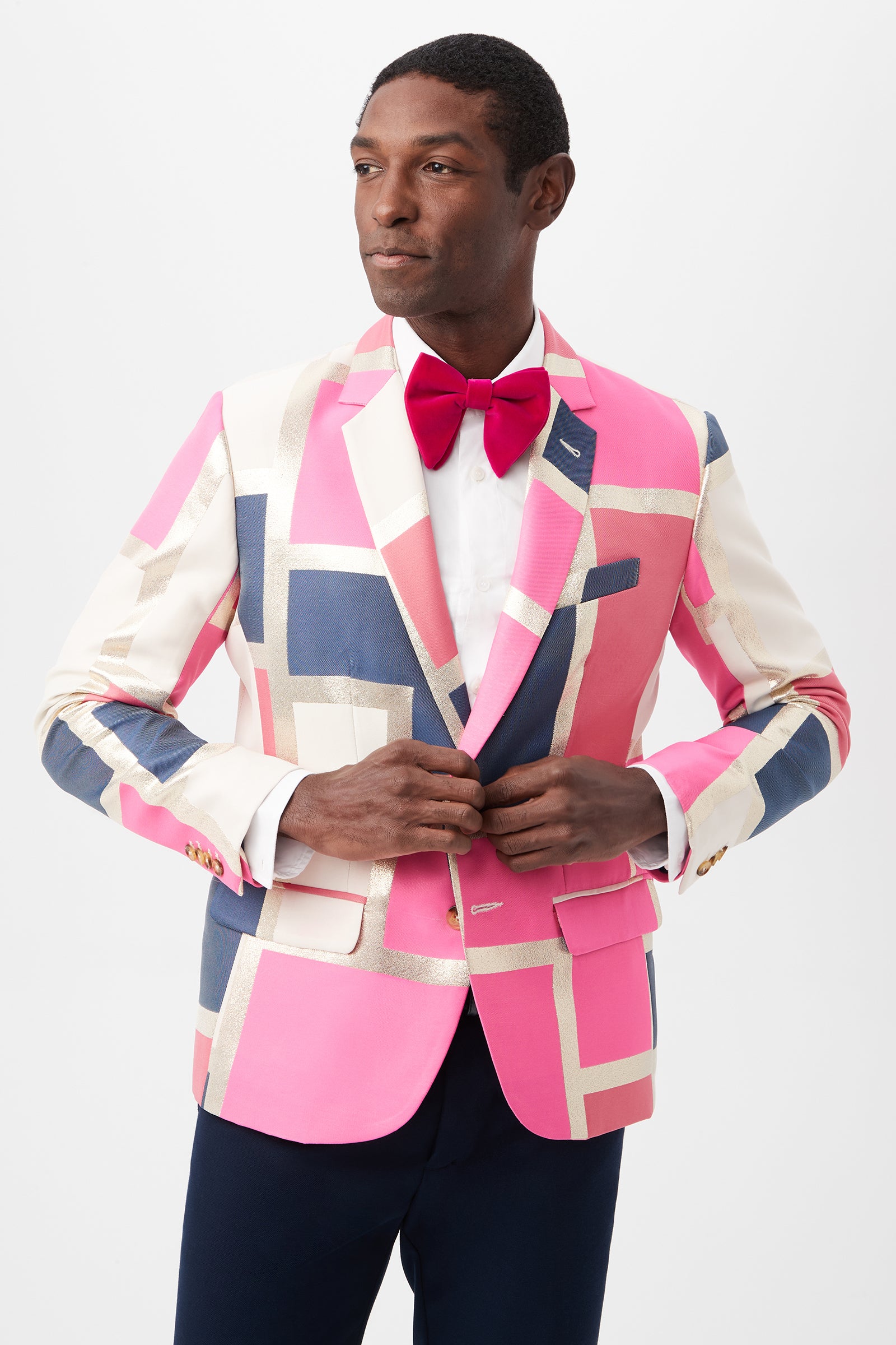 A man stands buttoning the Thurston Blazer - Block Party Jacquard in Multi over a white shirt, red bow tie, and dark pants against a plain white background.