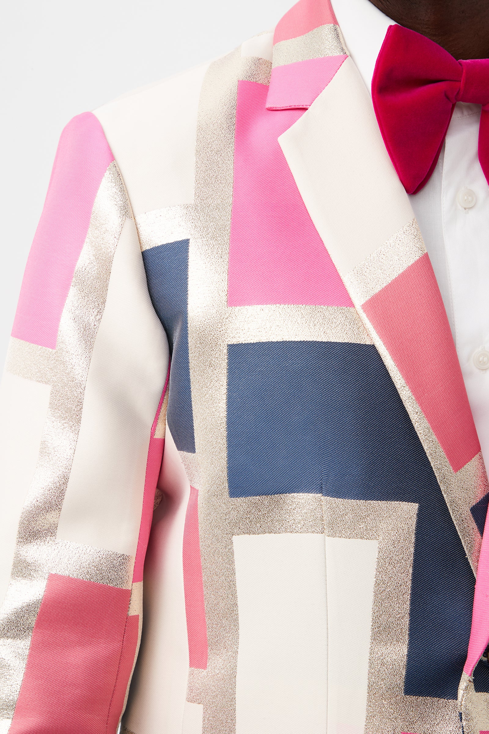 Close-up of a person wearing the Thurston Blazer - Block Party Jacquard, a colorful suit jacket with geometric pink, cream, gold glitter, and navy blue patterns, paired with a white shirt and bright pink bow tie.