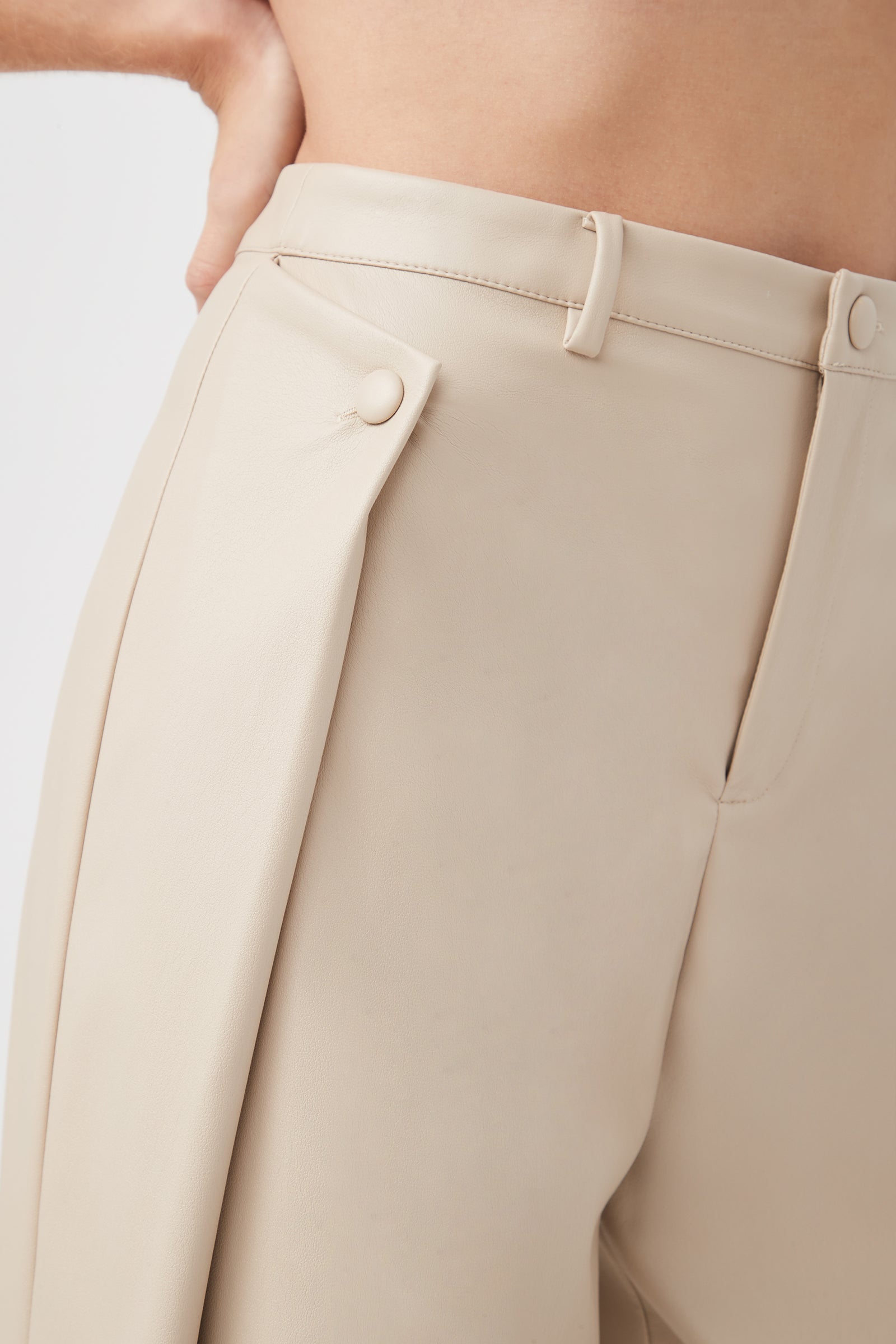 Close-up of a person wearing the Bexley Pant, a high-waisted beige style featuring a decorative side flap with button detail. The person’s hand rests on their hip, and the upper torso is visible.