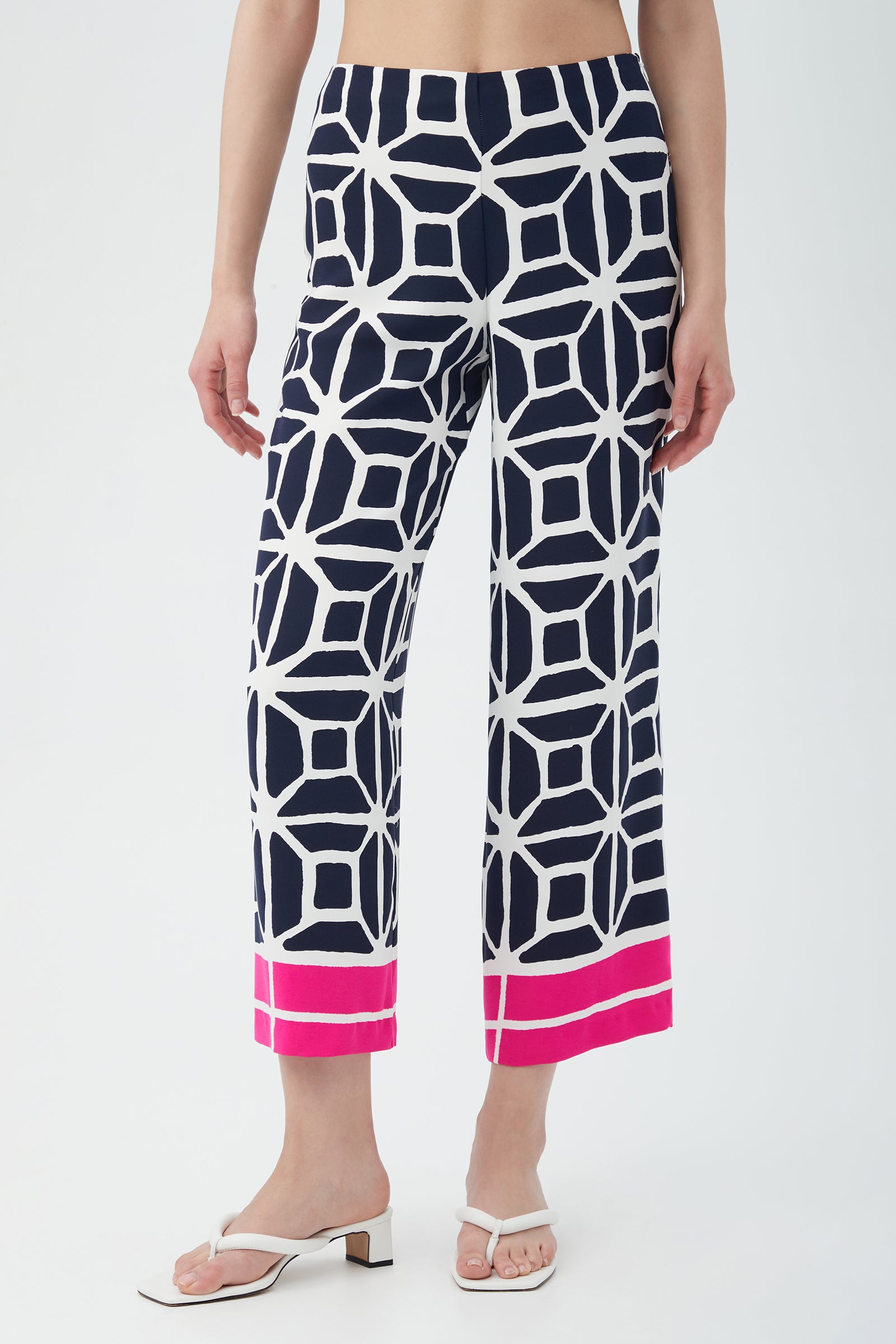 A woman models the Orlett Wide-Leg Crop Pant - Durell Georgette in a multi-color geometric pattern with a pink hem, paired with white sandals. The image is waist-down.