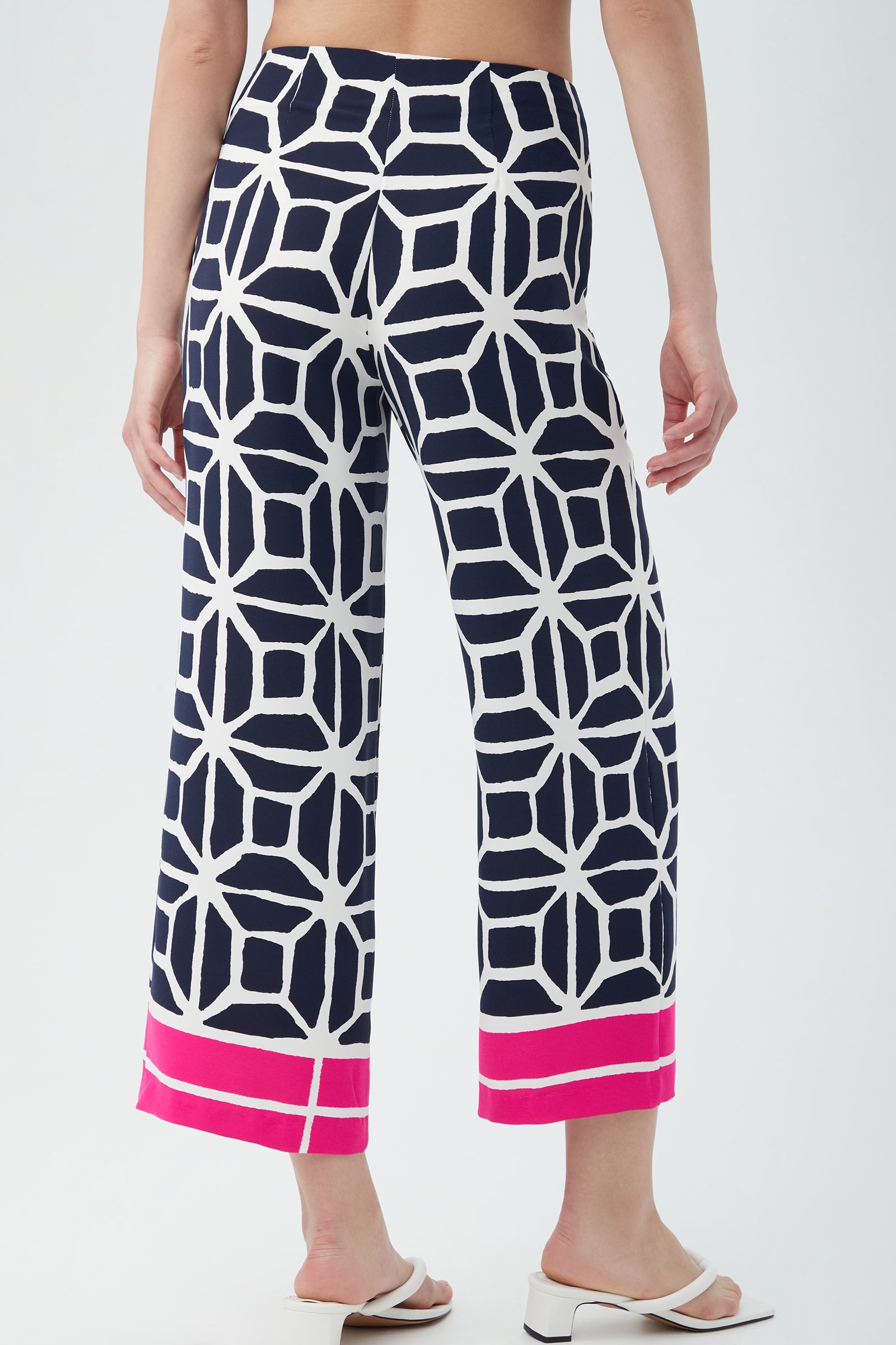 A person is seen from the back wearing Orlett Wide-Leg Crop Pant - Durell Georgette, featuring a navy blue and white geometric pattern with bright pink hem bands, paired with white heeled sandals against a plain background.
