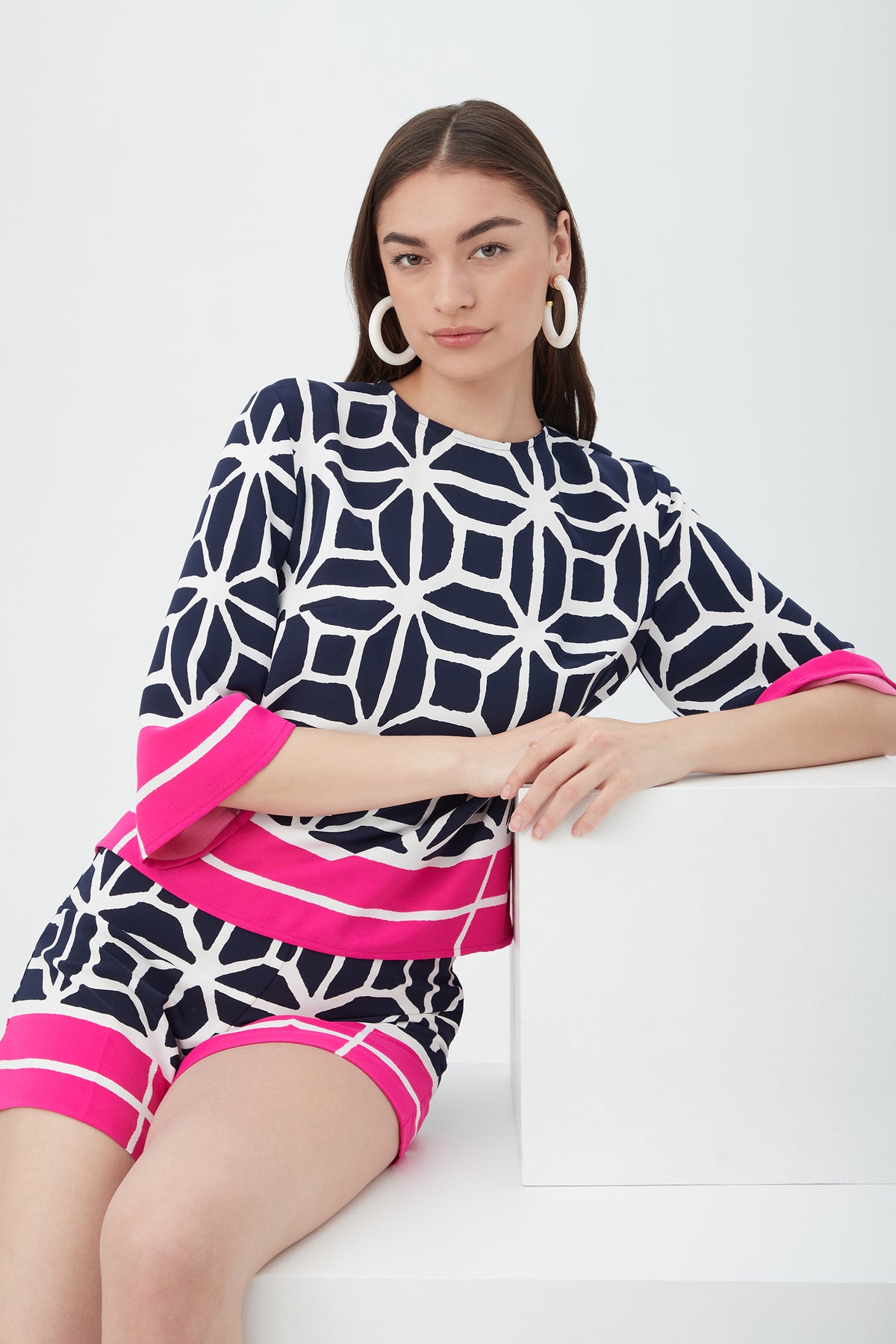 A woman with straight brown hair models the Shalyn Shorts - Durell Georgette in navy and white geometric print with bold pink stripes, paired with a matching top and large white hoop earrings, sitting confidently on a white bench.