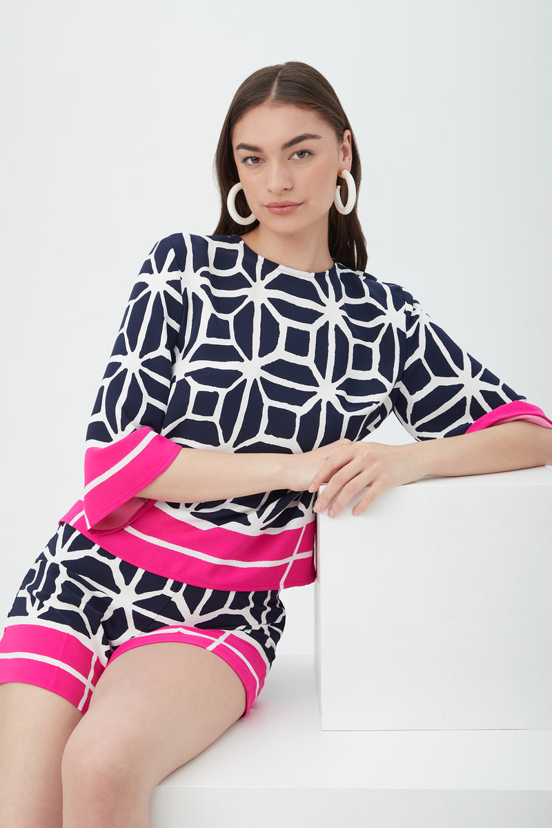 A woman with straight brown hair models the Shalyn Shorts - Durell Georgette in navy and white geometric print with bold pink stripes, paired with a matching top and large white hoop earrings, sitting confidently on a white bench.