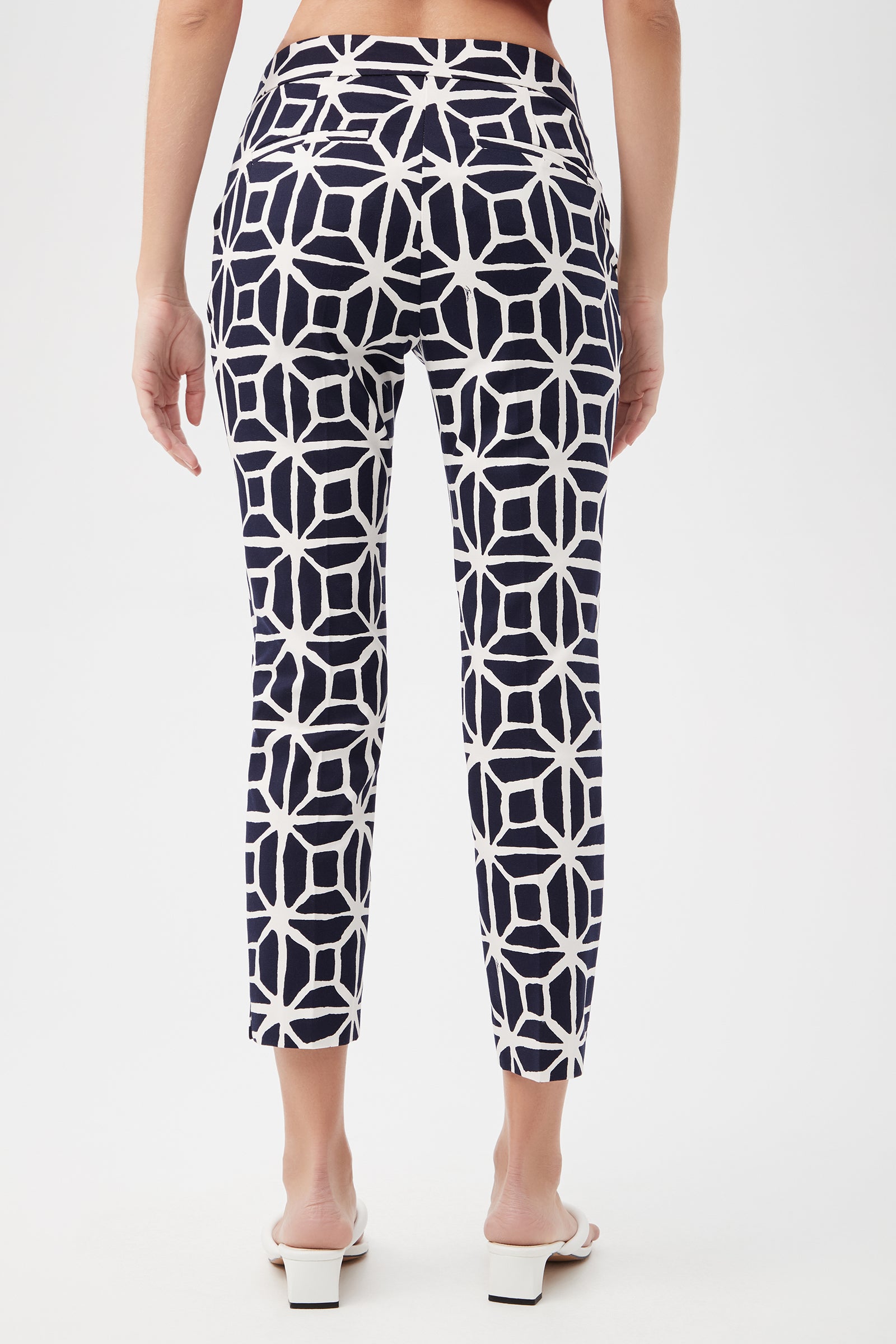 A woman faces away, wearing Moss 2 Slim Crop Pant - Durell Herringbone in navy with a white geometric pattern, paired with white open-back heeled sandals. The background is plain and light-colored.