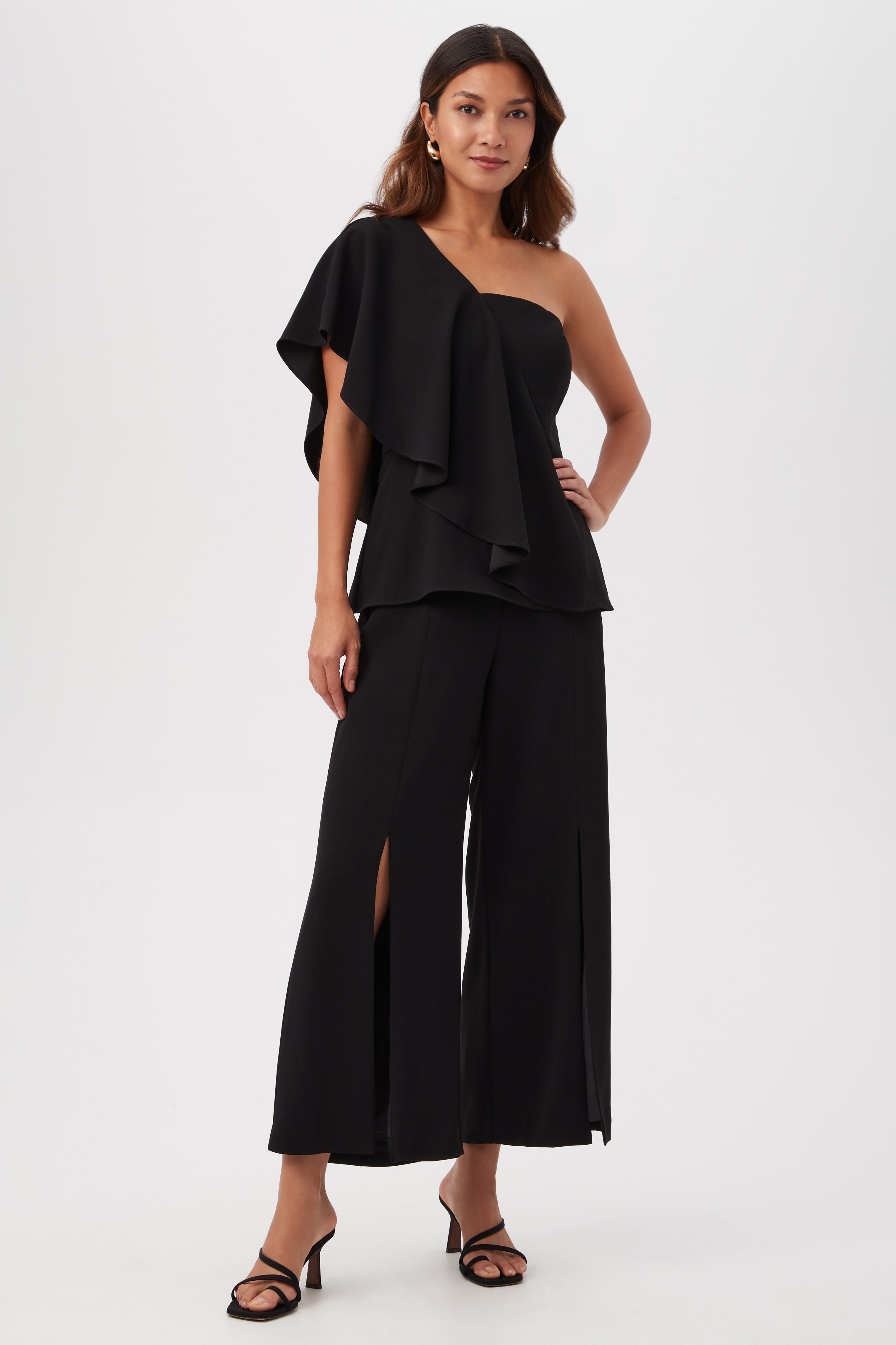A woman stands against a plain white background, wearing black Davon Cropped Wide-Leg Pants with front slits and a stylish one-shoulder ruffle top, paired with black strappy heels.