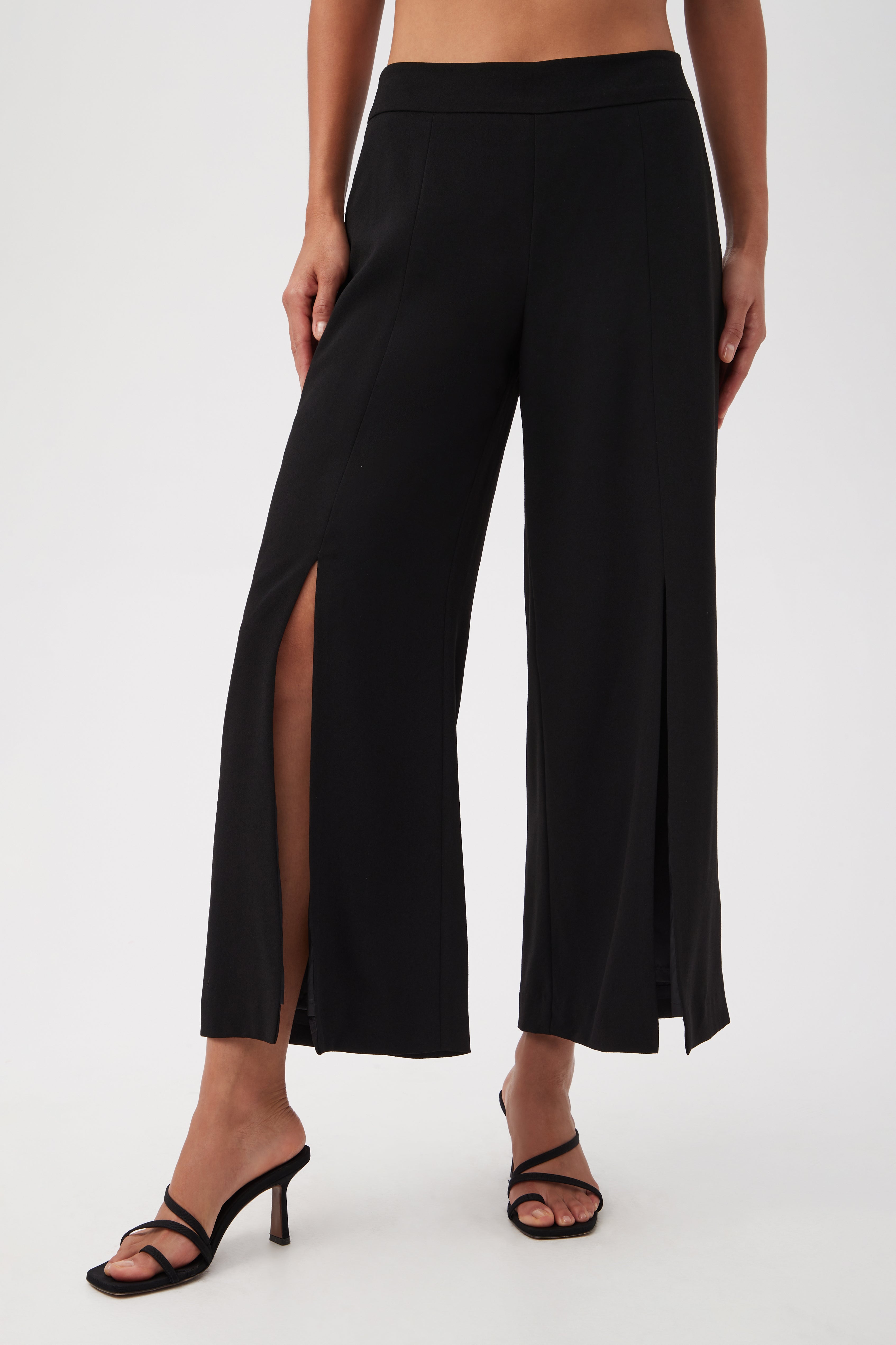 A person wears black Davon Cropped Wide-Leg Pants with front slits and black high-heeled sandals, standing against a plain white background. The image is cropped below the chest.