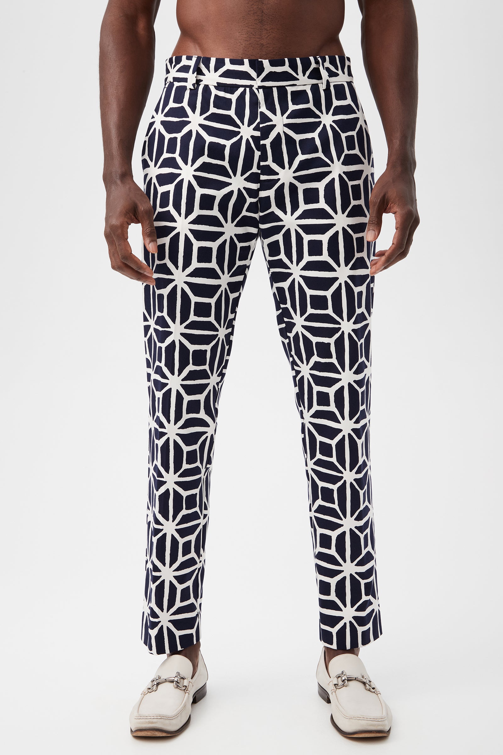 A person wearing the Clyde Slim Trouser - Durell Herringbone in indigo with a bold white geometric pattern and white loafers stands against a plain white background.