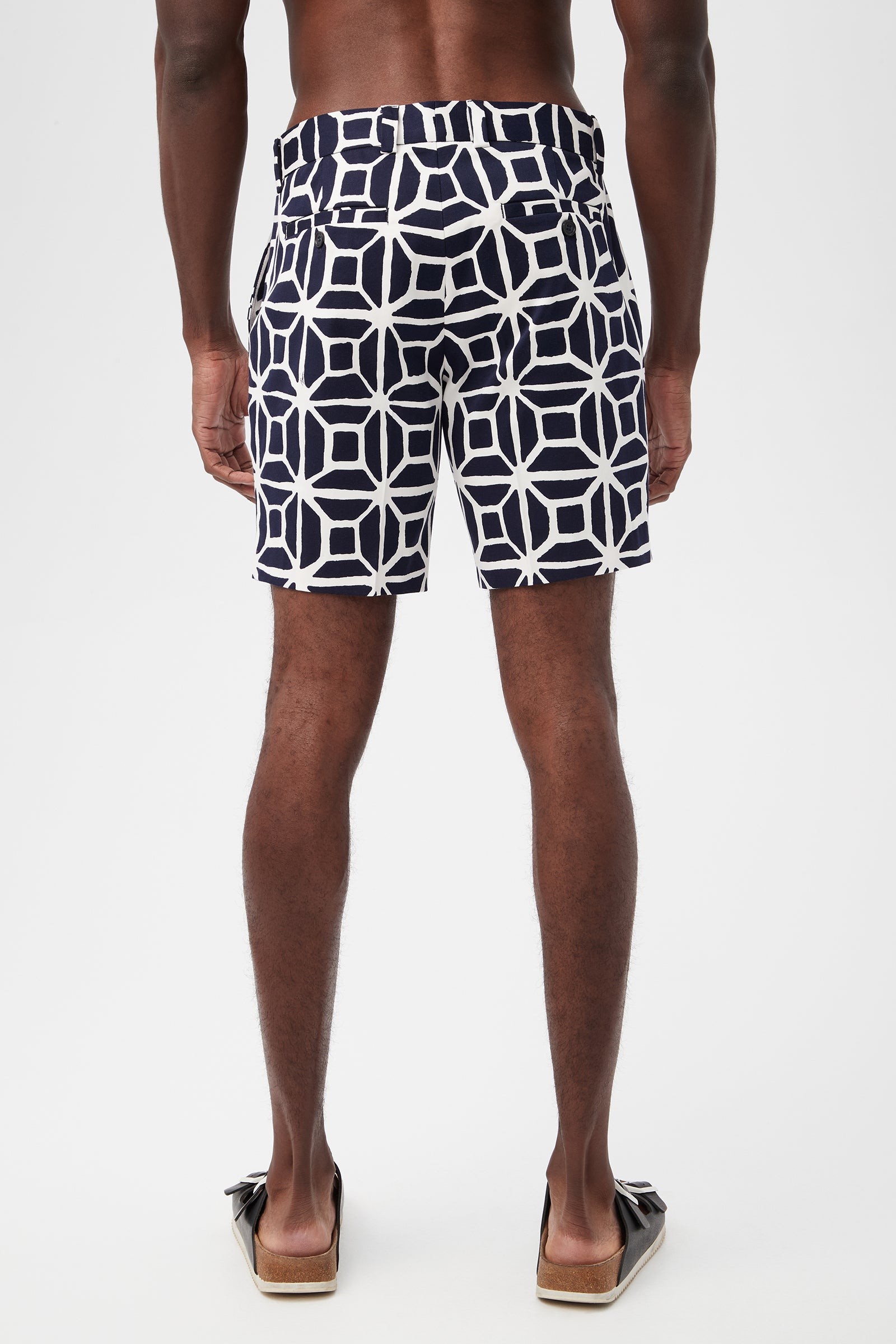 A man stands barefoot on a white background, shown from the back wearing Lawrence Short - Durell Herringbone swim trunks in navy with a bold white geometric print.