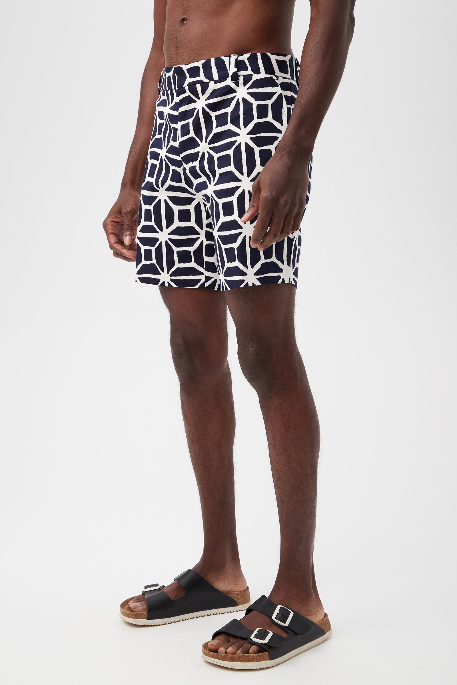 A man stands against a plain white background wearing the Lawrence Short - Durell Herringbone, featuring a navy blue and white geometric pattern. He is also wearing black buckle sandals and is bare-chested.