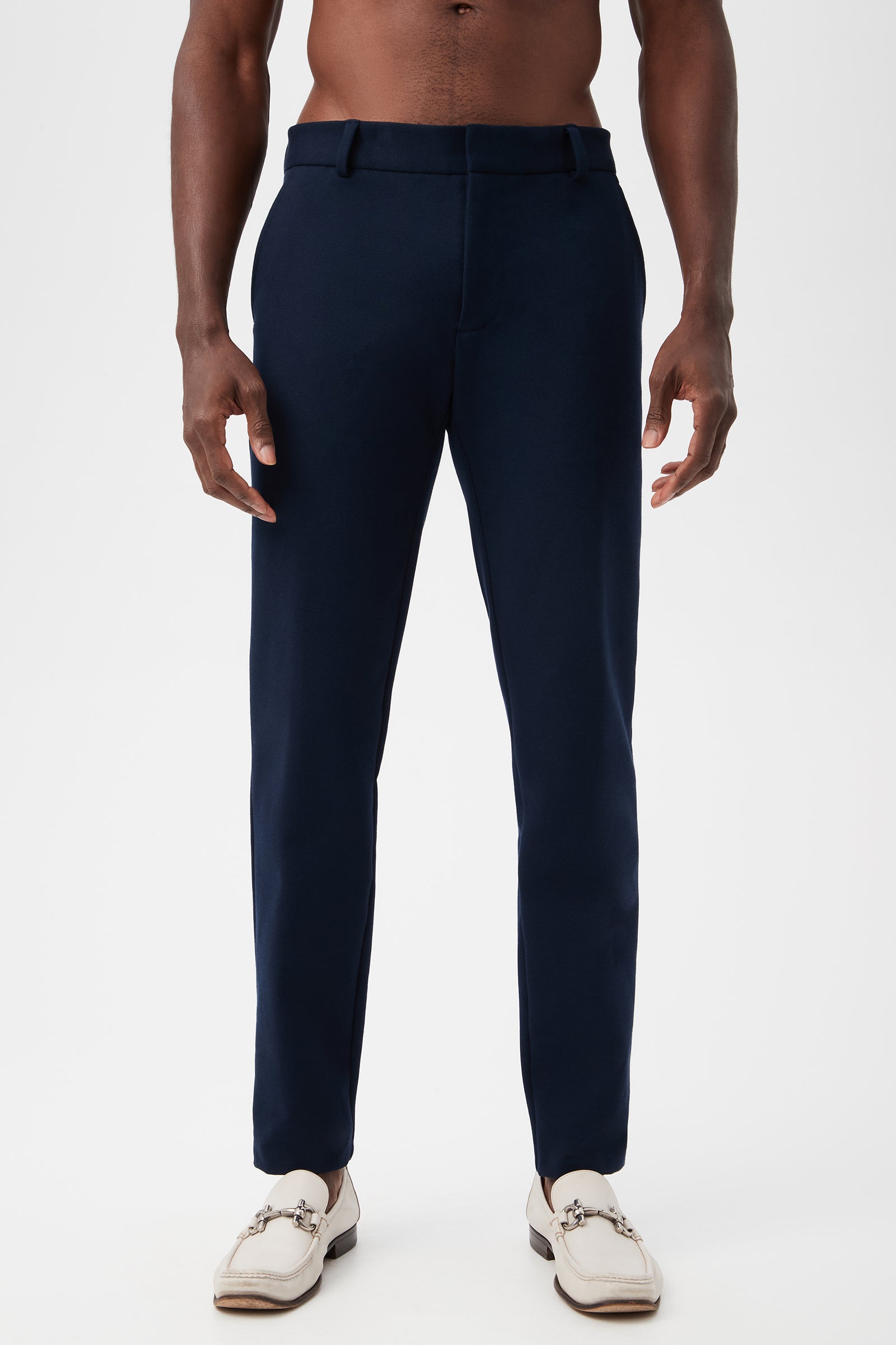 A man models the Clyde Slim Trouser - Stretch Piqué in indigo with white loafers, standing against a plain white background; his torso is bare and arms relaxed at his sides.