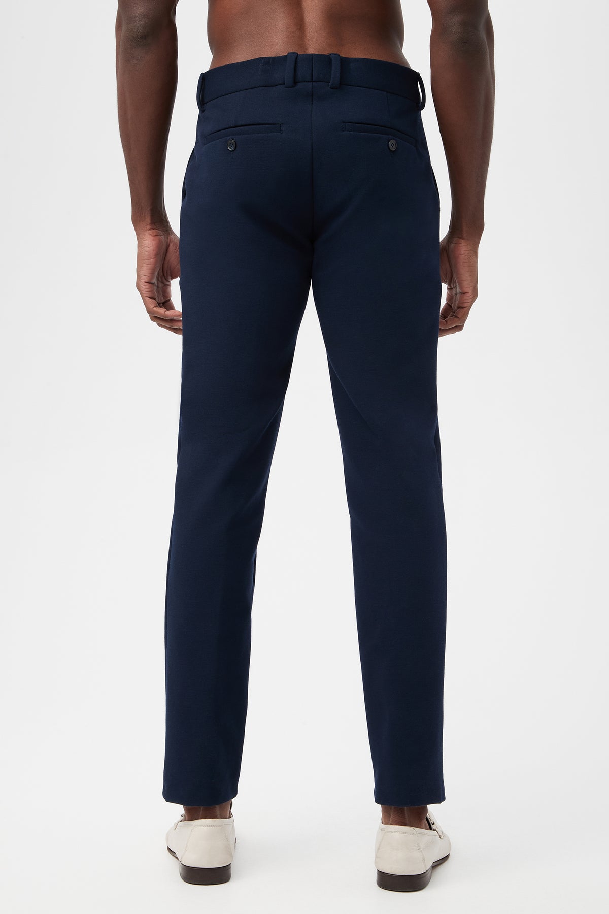 A man is seen from the back wearing the Clyde Slim Trouser - Stretch Piqué in navy blue, featuring belt loops, side pockets, and two buttoned back pockets. He pairs them with white shoes against a plain white background.
