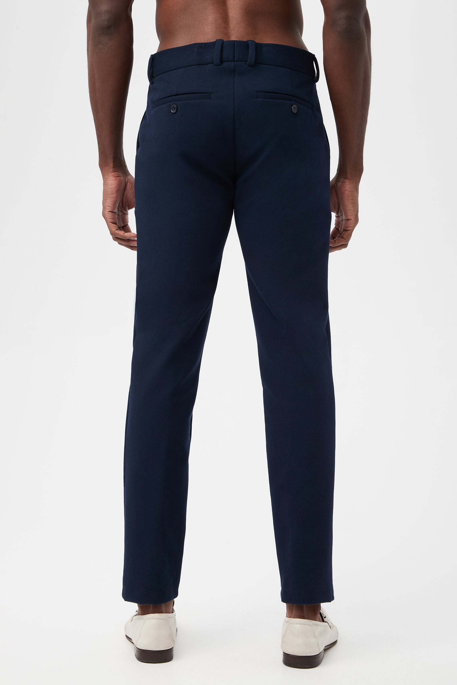 A man is seen from the back wearing the Clyde Slim Trouser - Stretch Piqué in navy blue, featuring belt loops, side pockets, and two buttoned back pockets. He pairs them with white shoes against a plain white background.