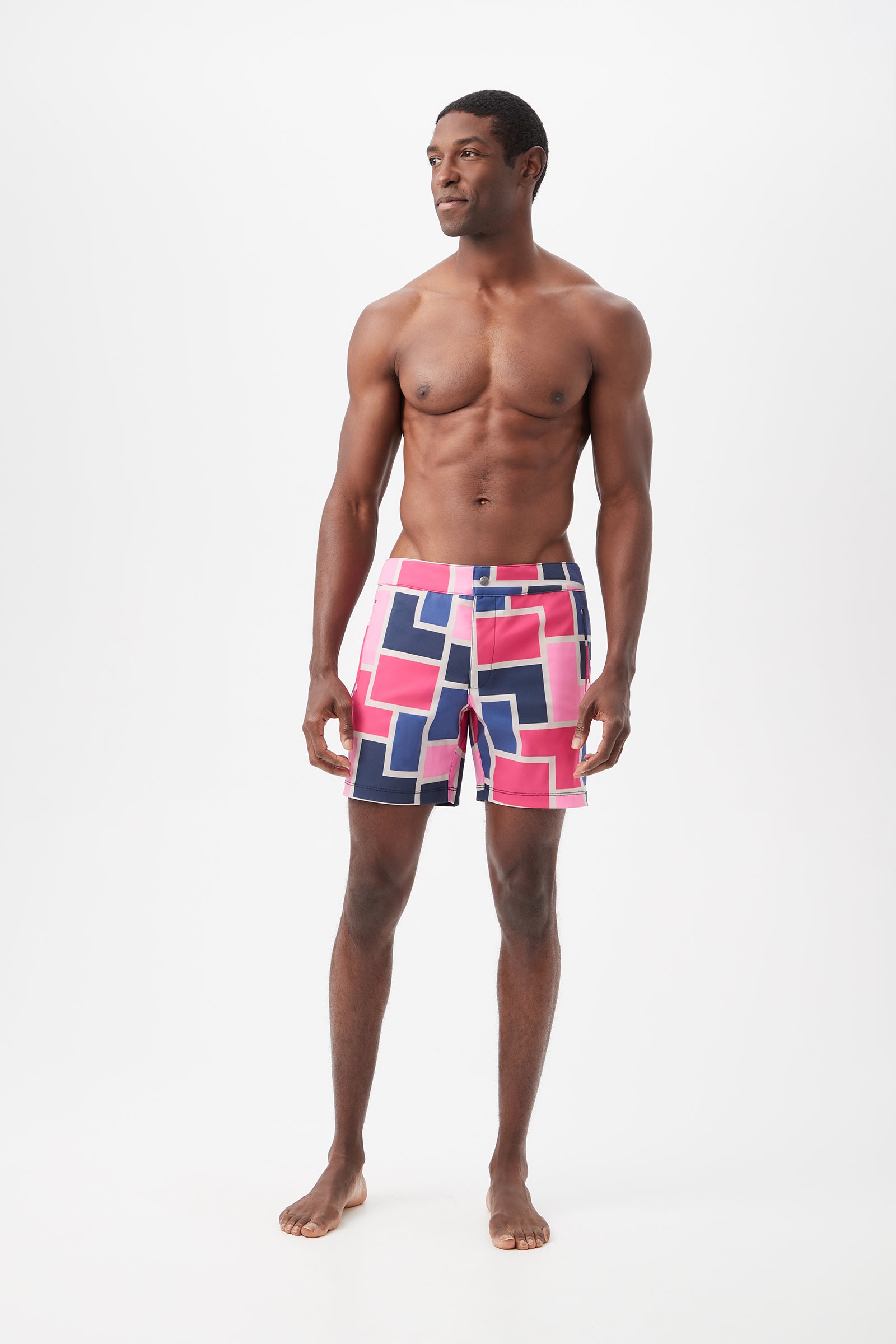 A man stands barefoot against a white background, wearing the Honolulu Board Short - Block Party, featuring colorful geometric patterns in pink, blue, and white. He is shirtless and gazes slightly to the side with a neutral expression.