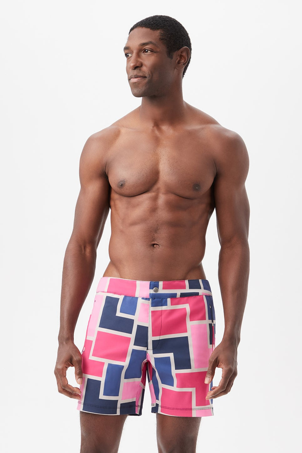 A fit, shirtless man stands confidently against a plain white background, wearing the Honolulu Board Short - Block Party in a multi-color geometric pattern.