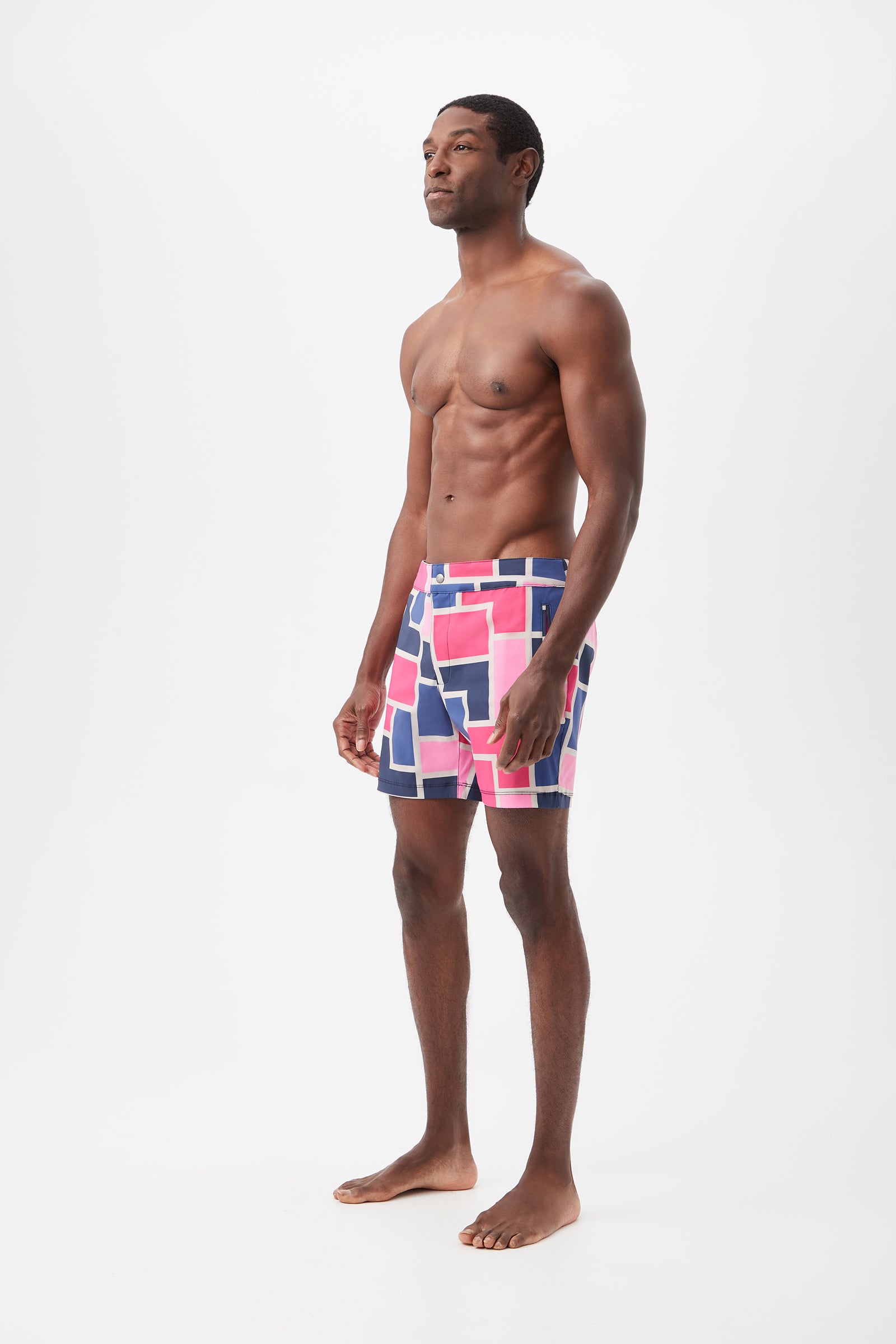 A man stands barefoot against a white background, wearing the Honolulu Board Short - Block Party featuring colorful geometric patterns in pink, blue, and white. He is shirtless, gazing slightly to the side with his arms relaxed at his sides.