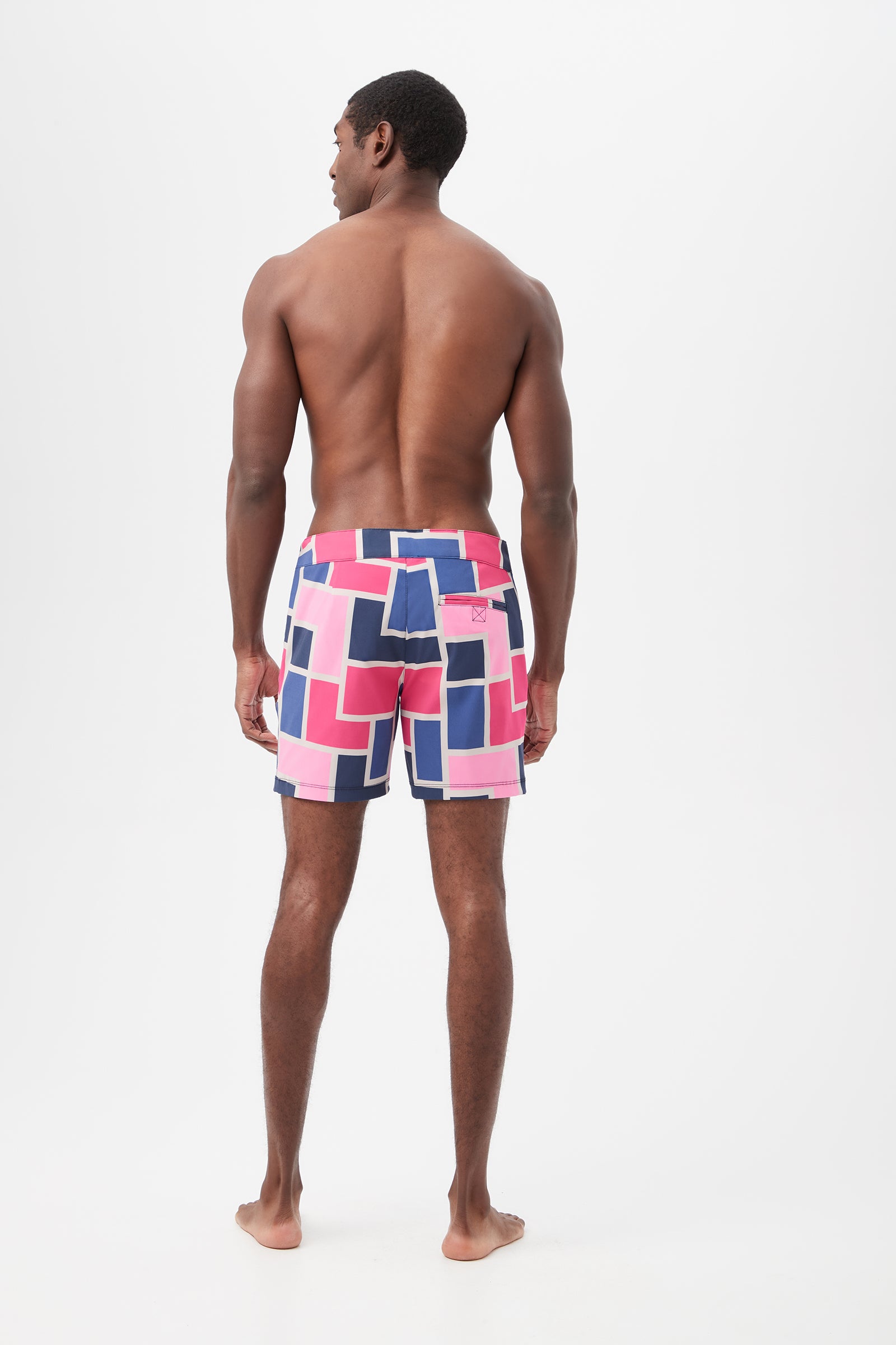 A man stands barefoot with his back to the camera, wearing the Honolulu Board Short - Block Party, featuring colorful geometric patterns in pink, blue, and white against a plain white background.