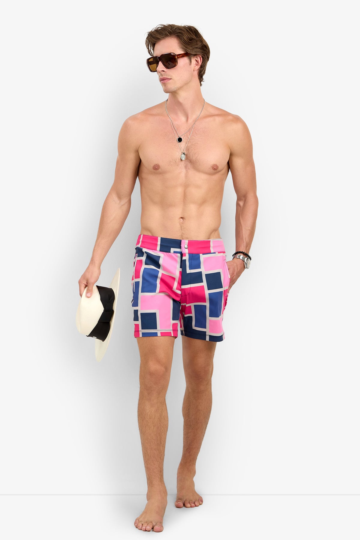 A shirtless man in sunglasses and Honolulu Boardshort holds a white hat while walking barefoot against a white background.