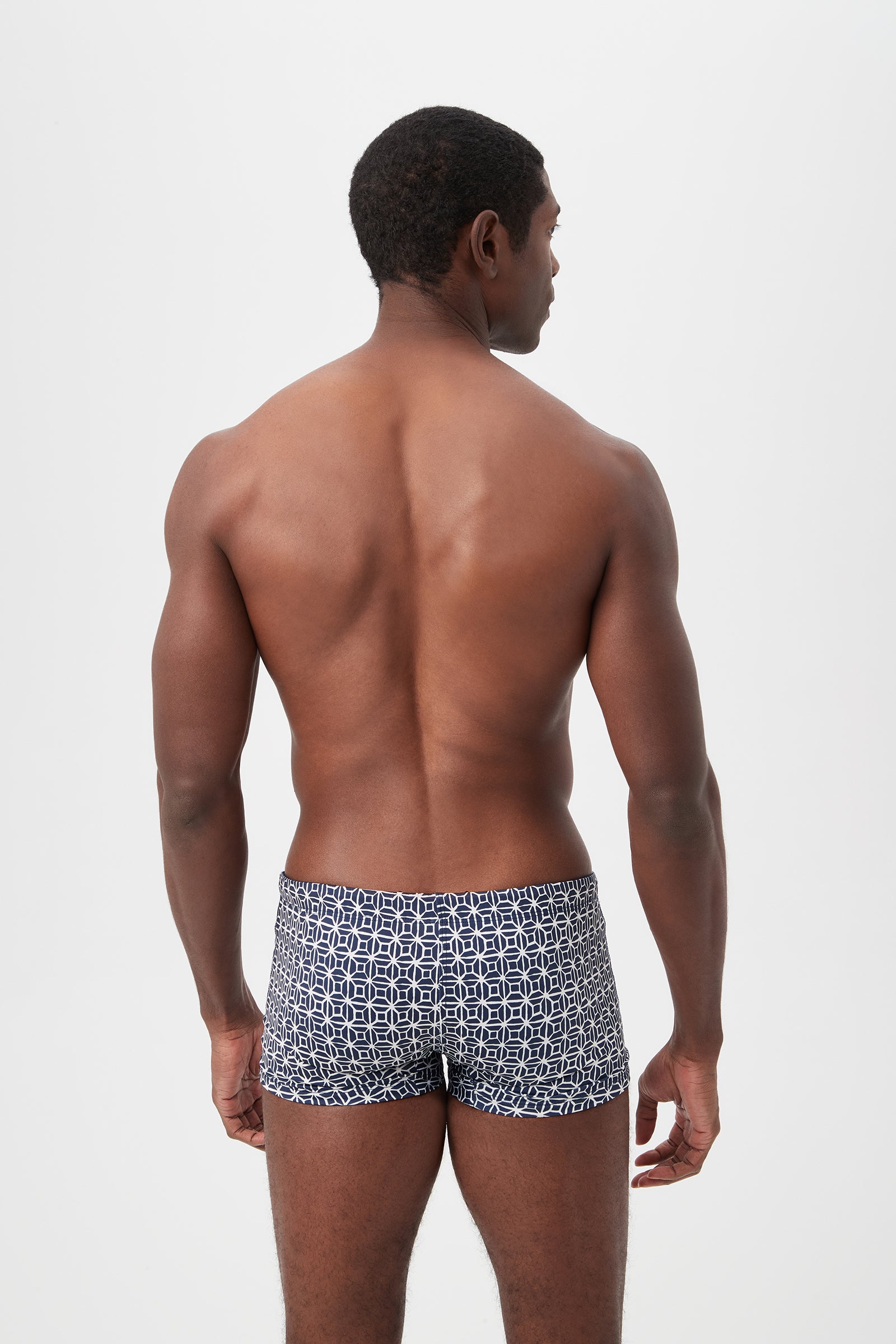 A man with short hair faces away, revealing his bare back. He is wearing the Seaside Reversible Swim Trunk in Durell Print, featuring a navy blue and white geometric pattern. The background is plain white.