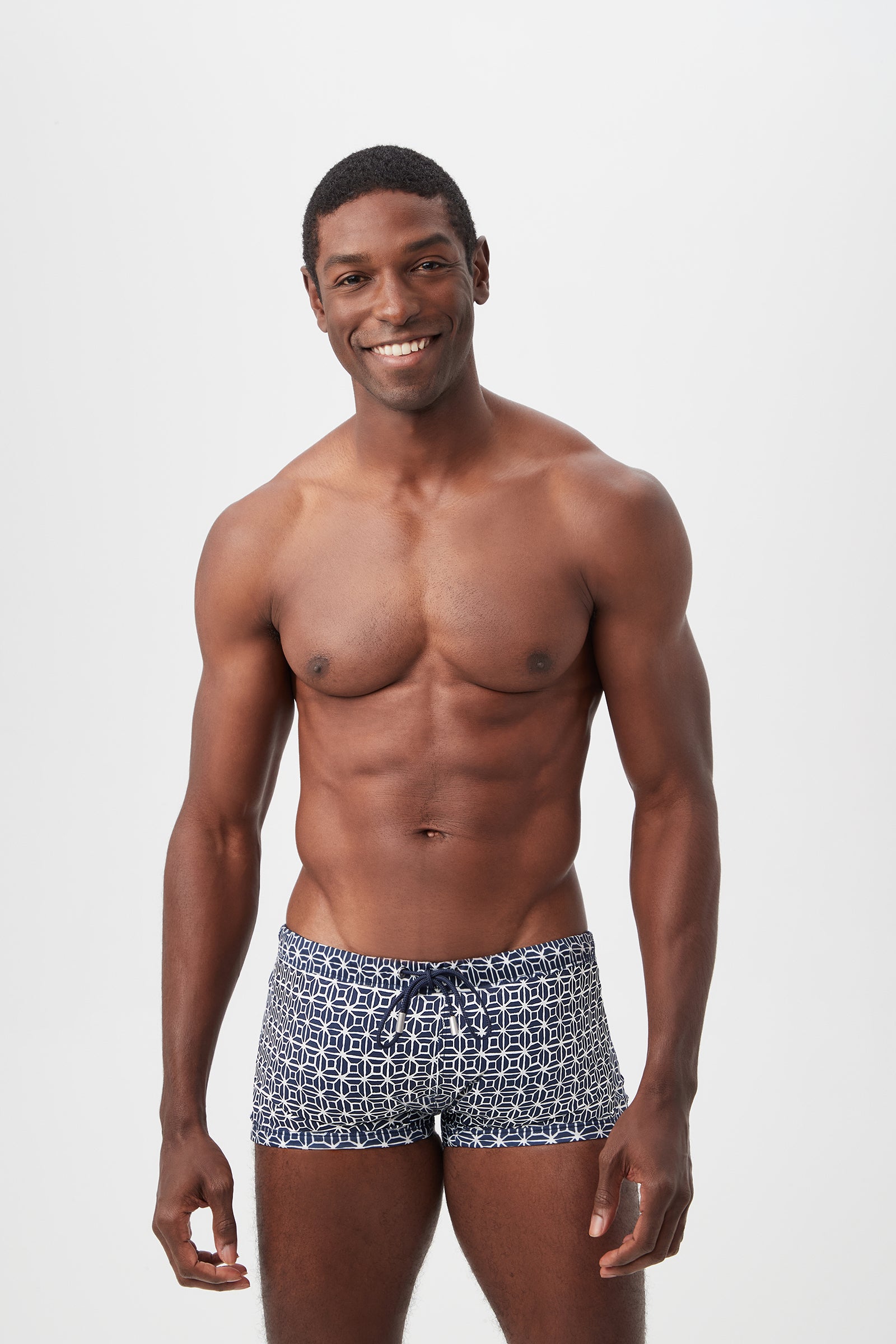 A smiling man stands against a plain white background, wearing Seaside Reversible Swim Trunk in Durell Print, color Indigo Multi. He is shirtless, showing a fit and muscular physique.