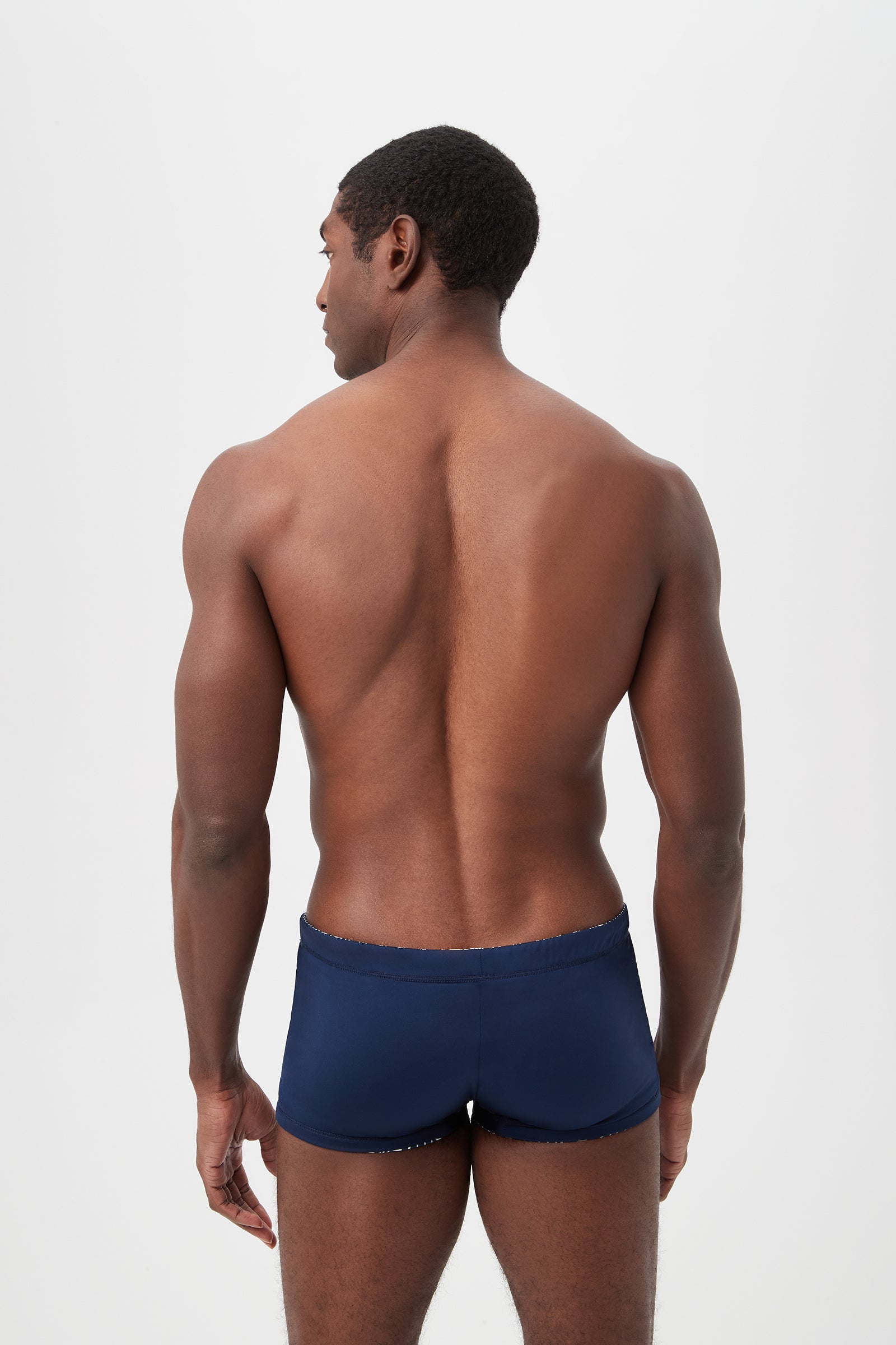 A muscular man, seen from behind against a plain light background, models the Seaside Reversible Swim Trunk in Durell Print.
