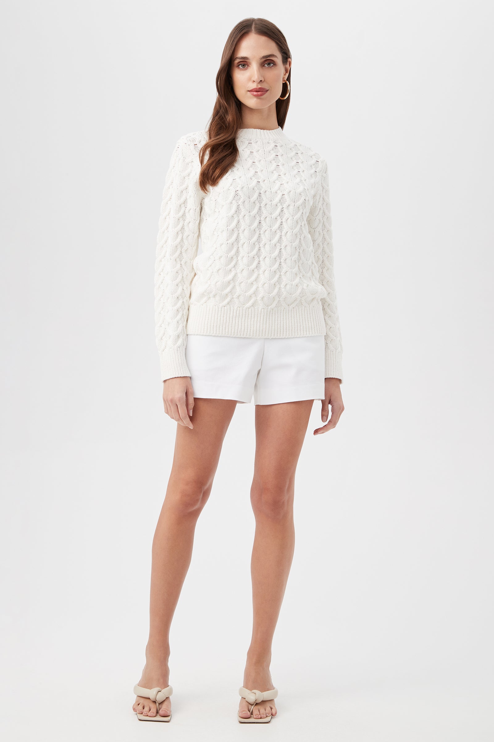 A woman with long brown hair and a neutral expression stands against a white background, wearing the Corazon Heart Knit Crewneck Sweater, white shorts, and beige heeled sandals.