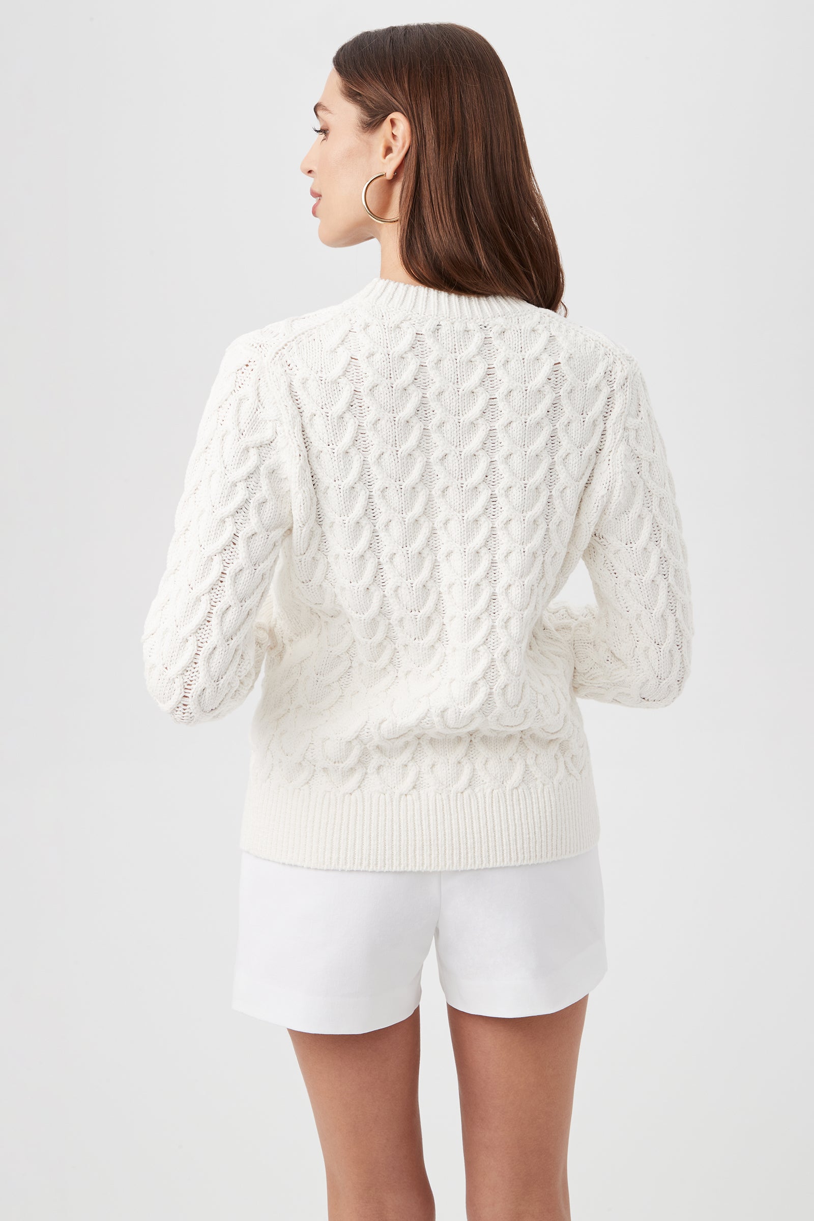 A woman with straight brown hair is seen from the back, wearing the Corazon Heart Knit Crewneck Sweater in white and matching shorts, one hand on her hip, standing against a plain light background.
