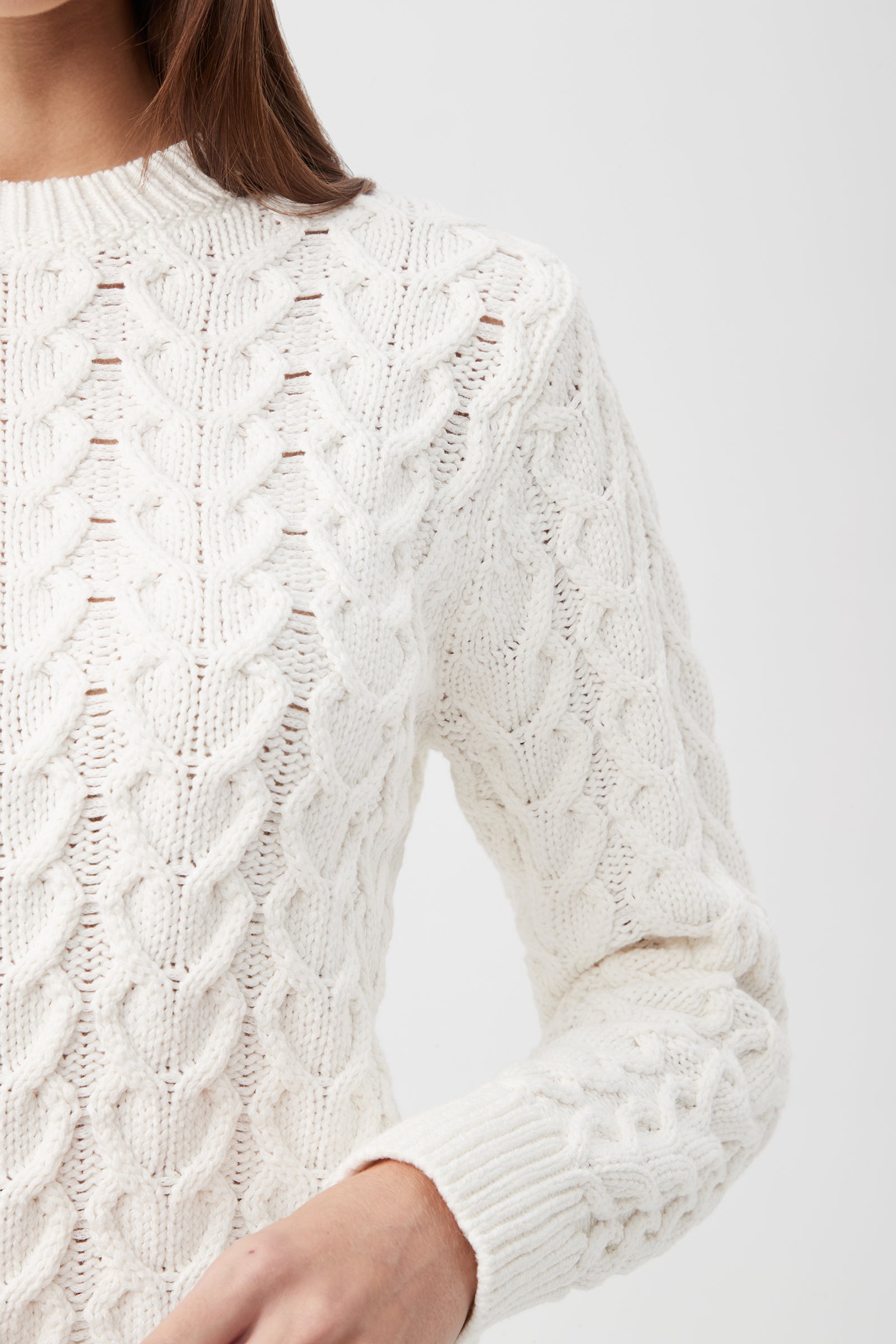 A person wearing the Corazon Heart Knit Crewneck Sweater in white, featuring long sleeves and a cable-knit design with a high neckline, shown from shoulders to waist against a plain light background.