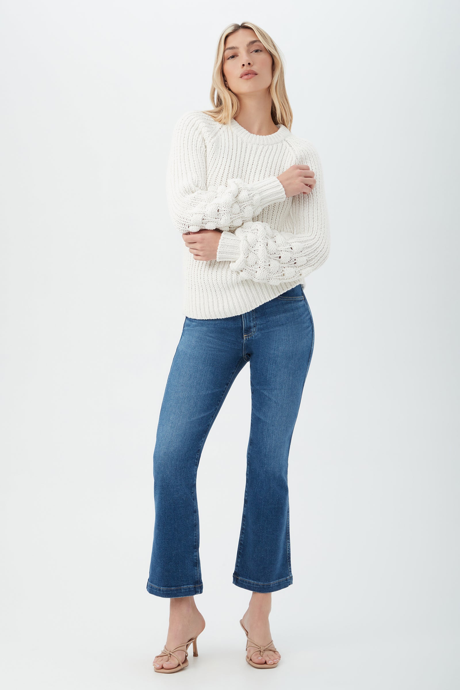 Wearing the Bellanca 2 Sweater, a white textured knit with decorative sleeves, paired with blue flared jeans and beige open-toe heels, a person poses confidently against a plain white background.