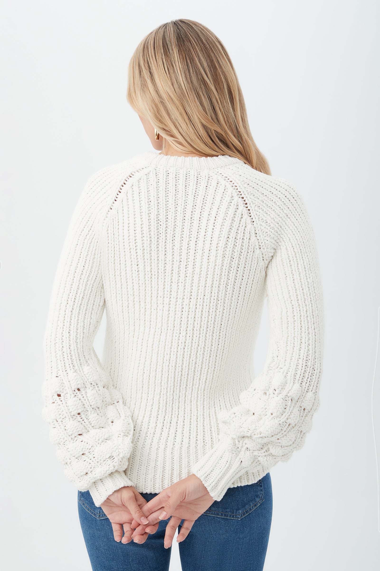 A woman with long blonde hair, seen from behind, wears the Bellanca 2 Sweater—a white knit with textured, bubble-like sleeves—paired with blue jeans. She stands against a plain background, hands clasped behind her back.