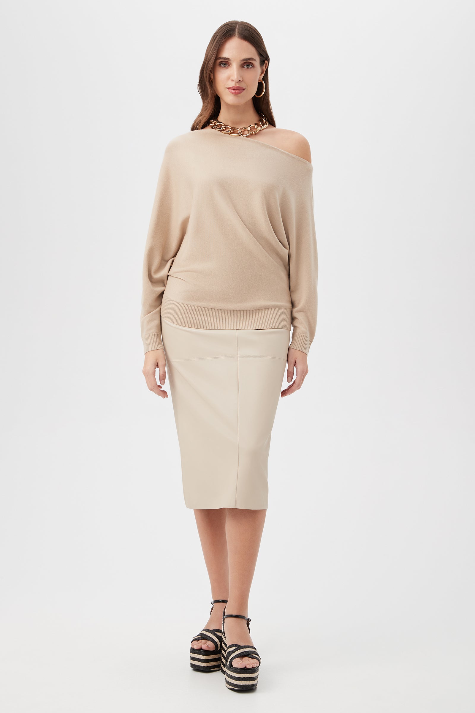 A woman with long brown hair and a neutral expression stands against a white background, wearing the Montage Sweater in beige, a light beige knee-length pencil skirt, black platform sandals, and a chunky gold necklace.