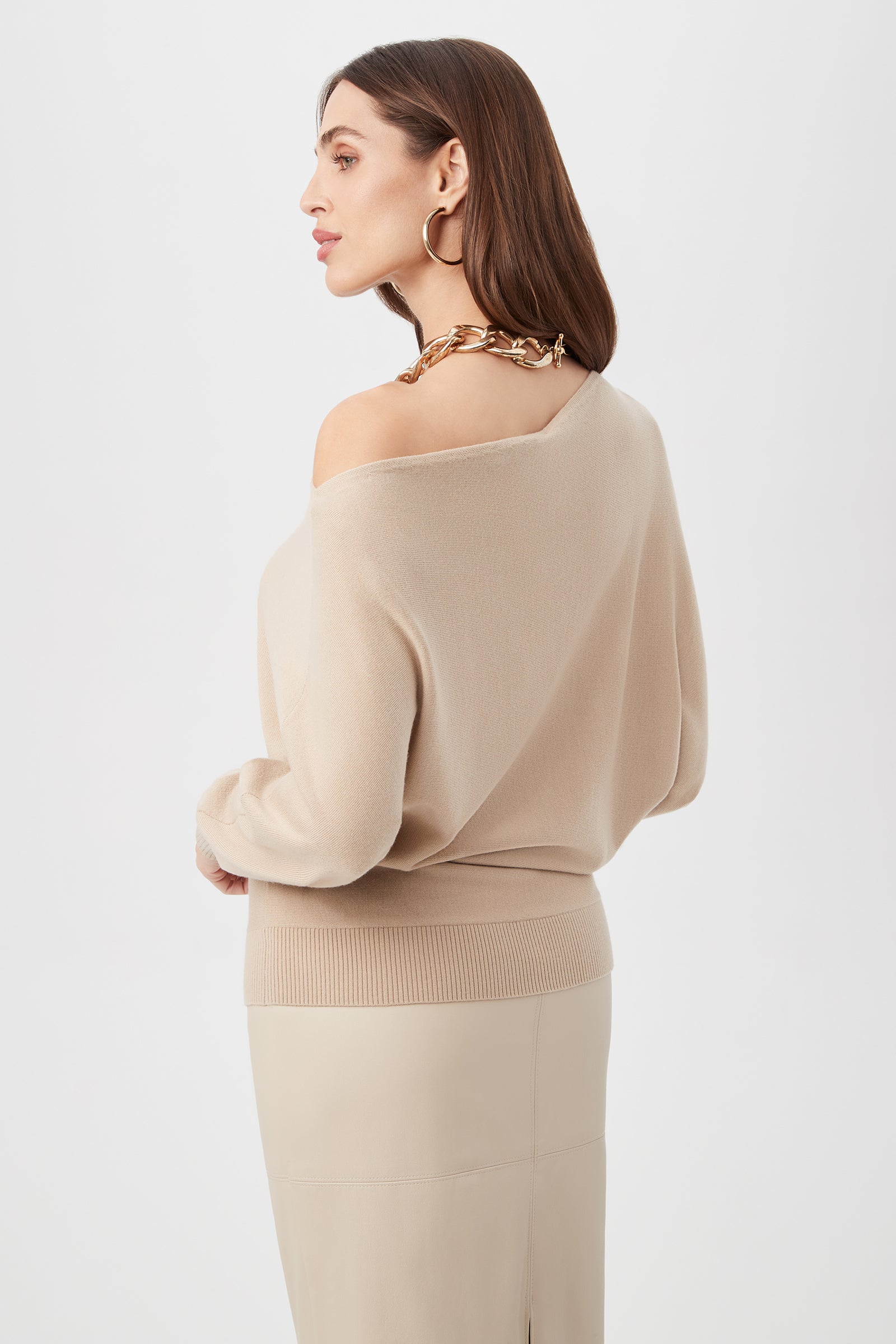A woman with straight brown hair wears the Montage Sweater in beige, paired with a gold chunky chain necklace and beige skirt, standing against a plain white background.