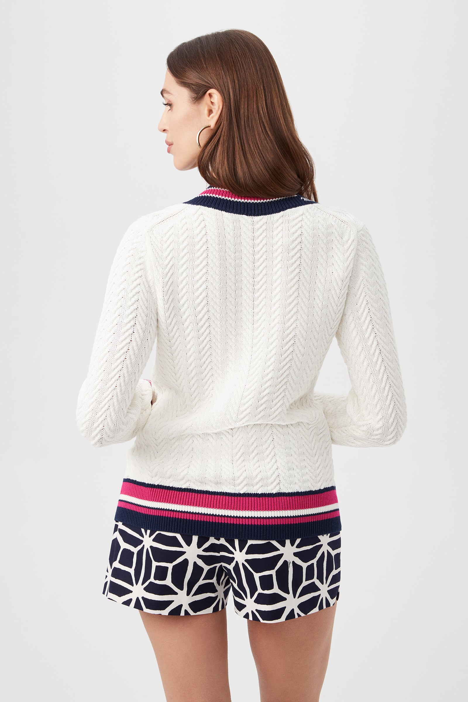 A woman with brown hair, seen from behind, wears the Seapointe Sweater—white and textured with navy, pink, and white collar and cuffs—paired with navy shorts featuring a white geometric pattern.