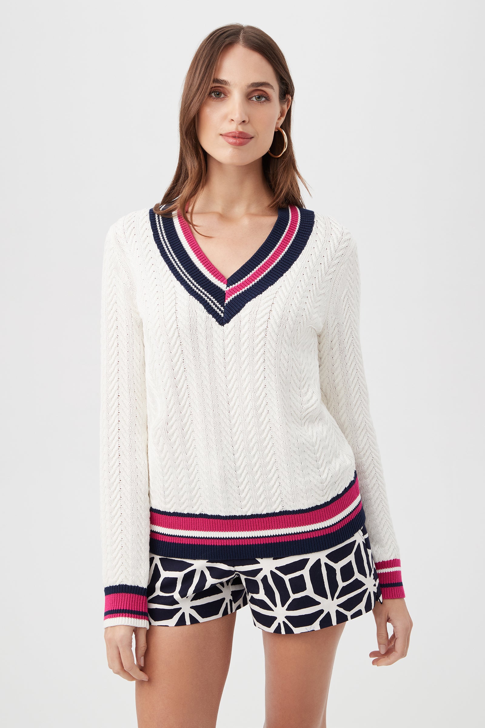 A woman wears the Seapointe Sweater in multi, featuring a white V-neck with navy and pink stripes, paired with patterned navy and white shorts, standing against a plain light background.