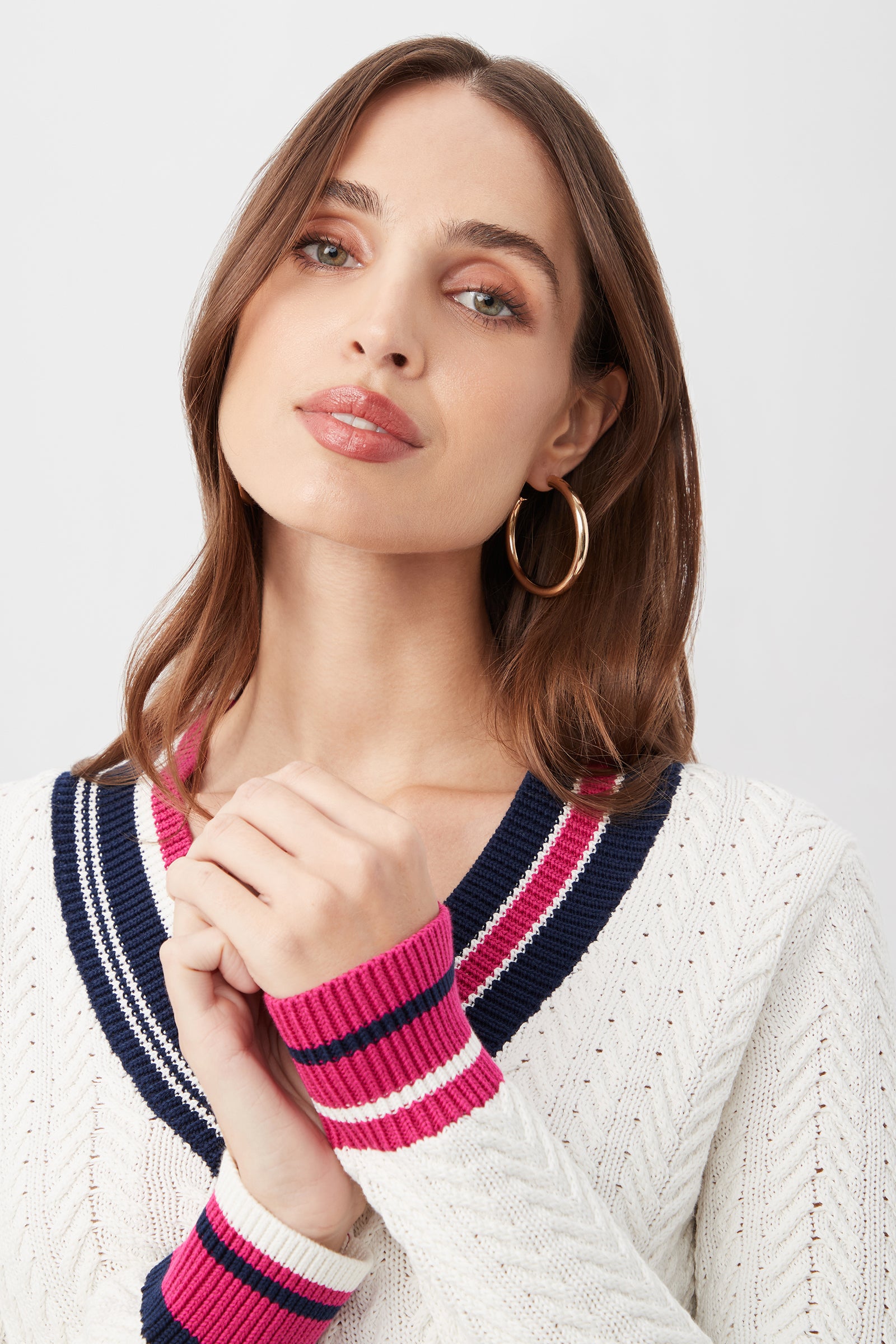 A woman with light brown hair wears the Seapointe Sweater—a white knit with navy and pink striped trim—paired with large hoop earrings. She looks upward, hands gently clasped near her face, against a plain light background.