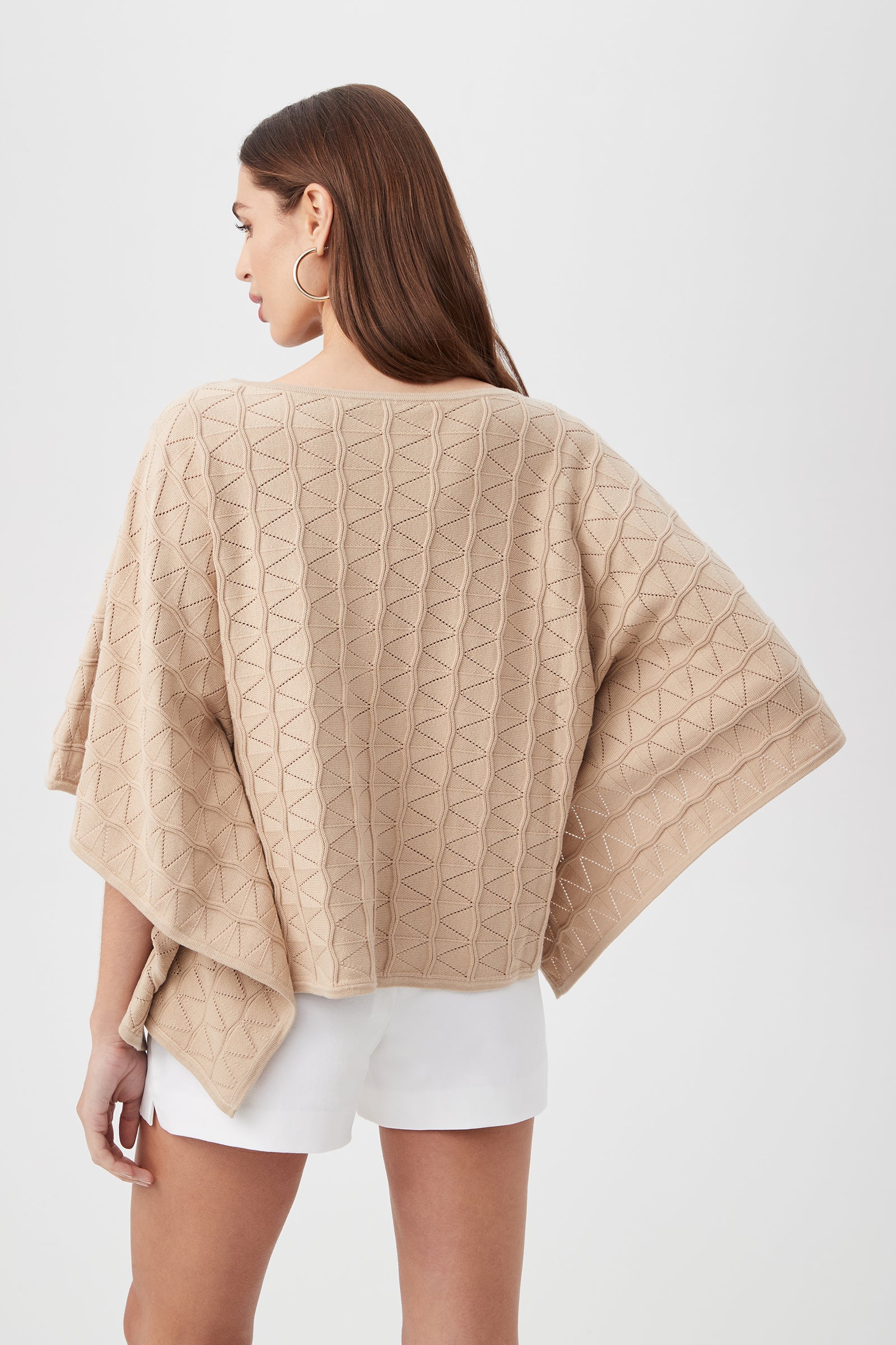 A woman with long brown hair, large hoop earrings, and white shorts models the Chic Wrap 2 Sweater—a beige textured poncho with a geometric pattern—while standing against a plain white background.