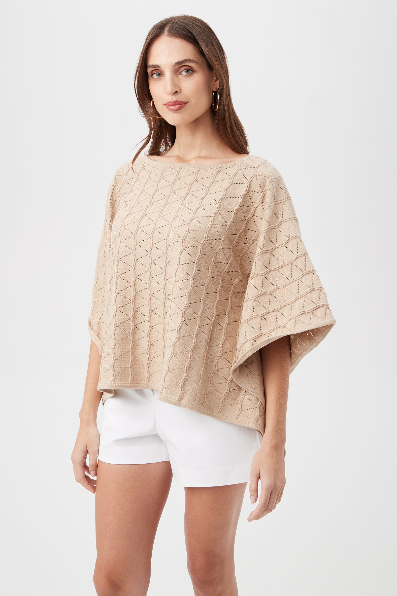A woman in the Chic Wrap 2 Sweater in chai, featuring a loose fit and geometric texture, pairs it with white shorts and poses against a plain white background. She has long brown hair and is turned slightly to the side.