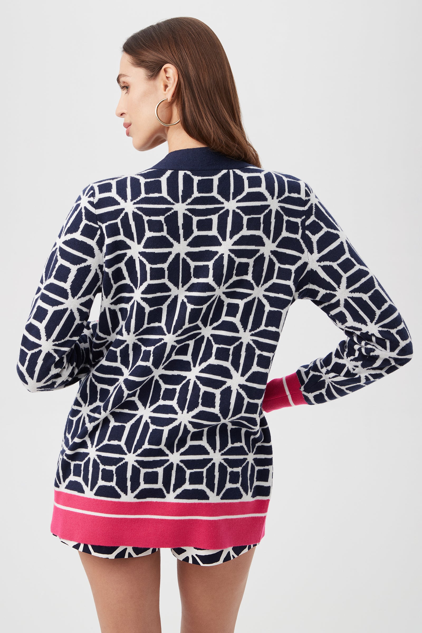 A woman with long brown hair and hoop earrings faces away, wearing the South Bay V-Neck Cardigan - Durell Knit in a navy and white geometric pattern with pink cuffs and hem, plus matching shorts, against a plain light background.