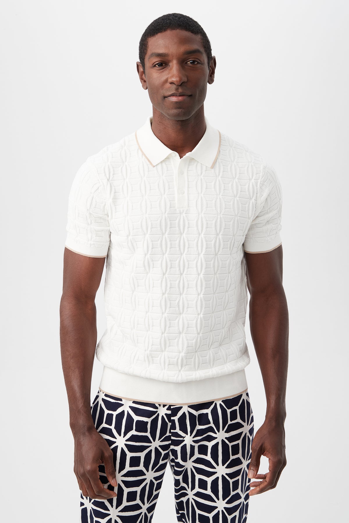 A man stands against a plain white background, wearing the Camilo Short-Sleeve Polo in Optic White and navy pants with a bold white geometric pattern. He faces forward with a neutral expression.