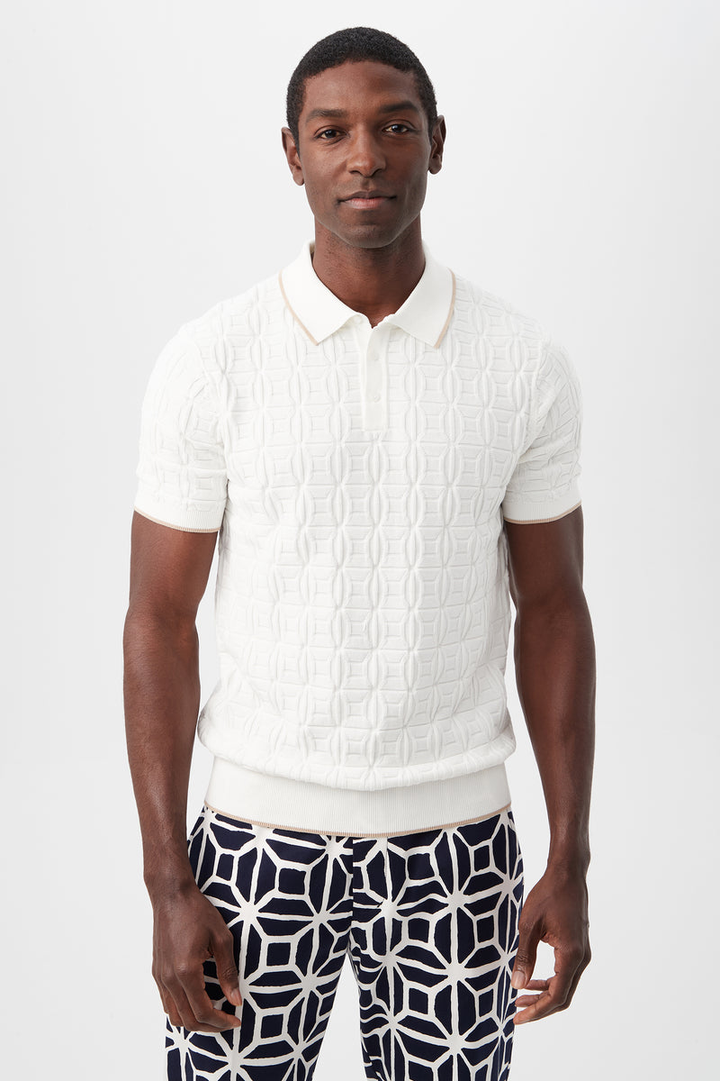 A man stands against a plain white background, wearing the Camilo Short-Sleeve Polo in Optic White and navy pants with a bold white geometric pattern. He faces forward with a neutral expression.