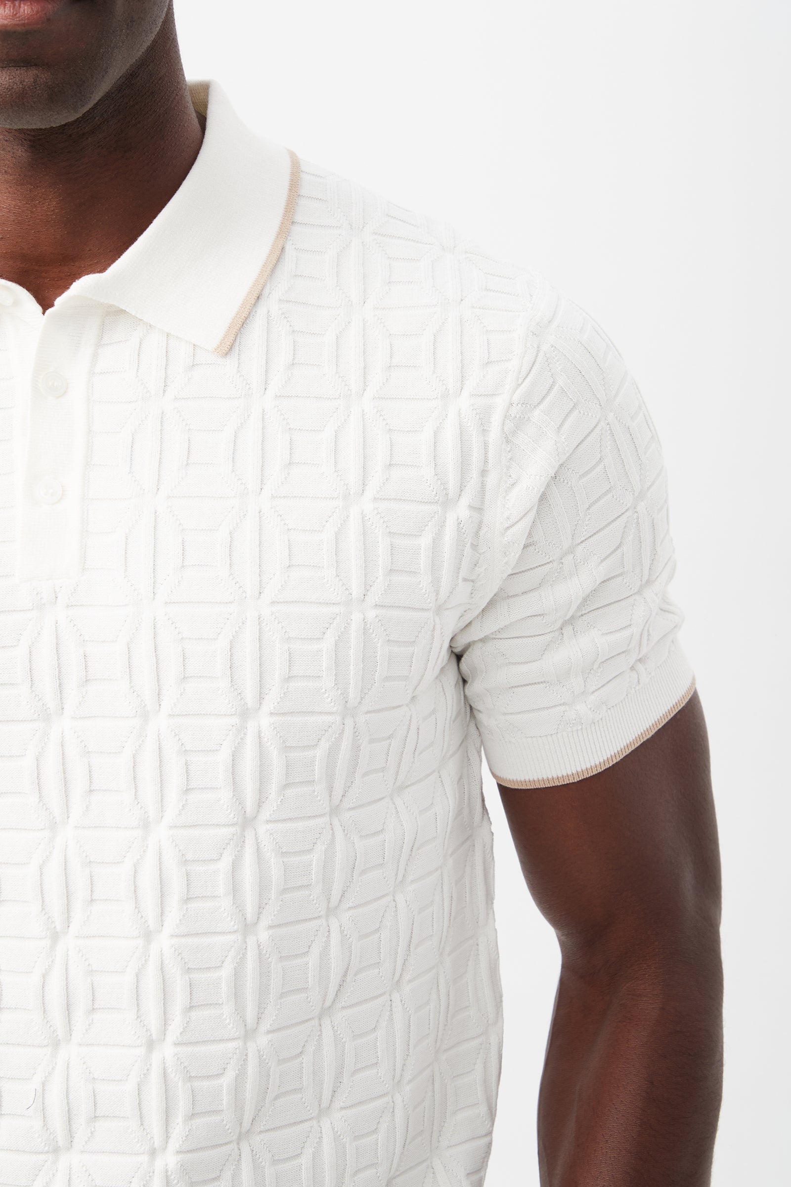 A man wears the Camilo Short-Sleeve Polo, featuring a textured white fabric, geometric pattern, beige collar trim, and white buttons, shown from shoulder to mid-torso.