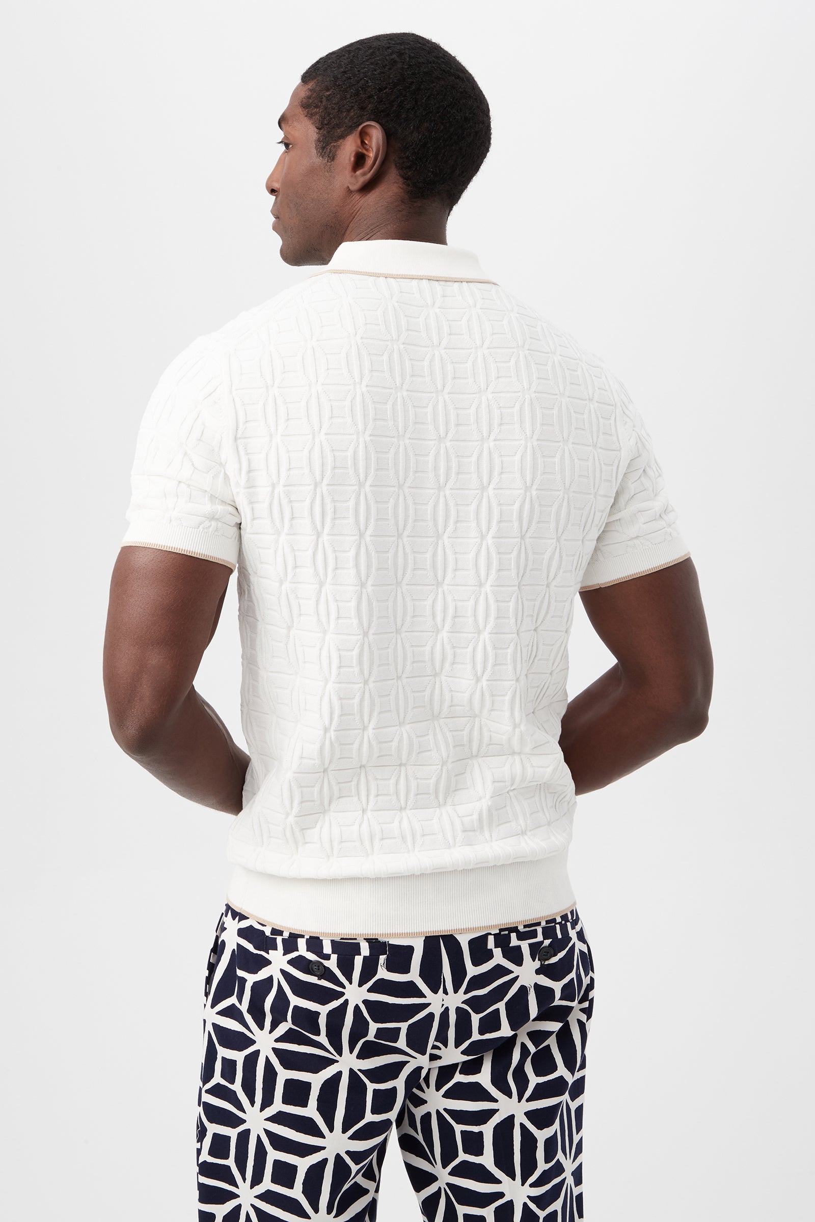 A man stands with his back turned, wearing the Camilo Short-Sleeve Polo—textured white with short sleeves—paired with navy and white geometric-patterned pants, set against a plain light background.