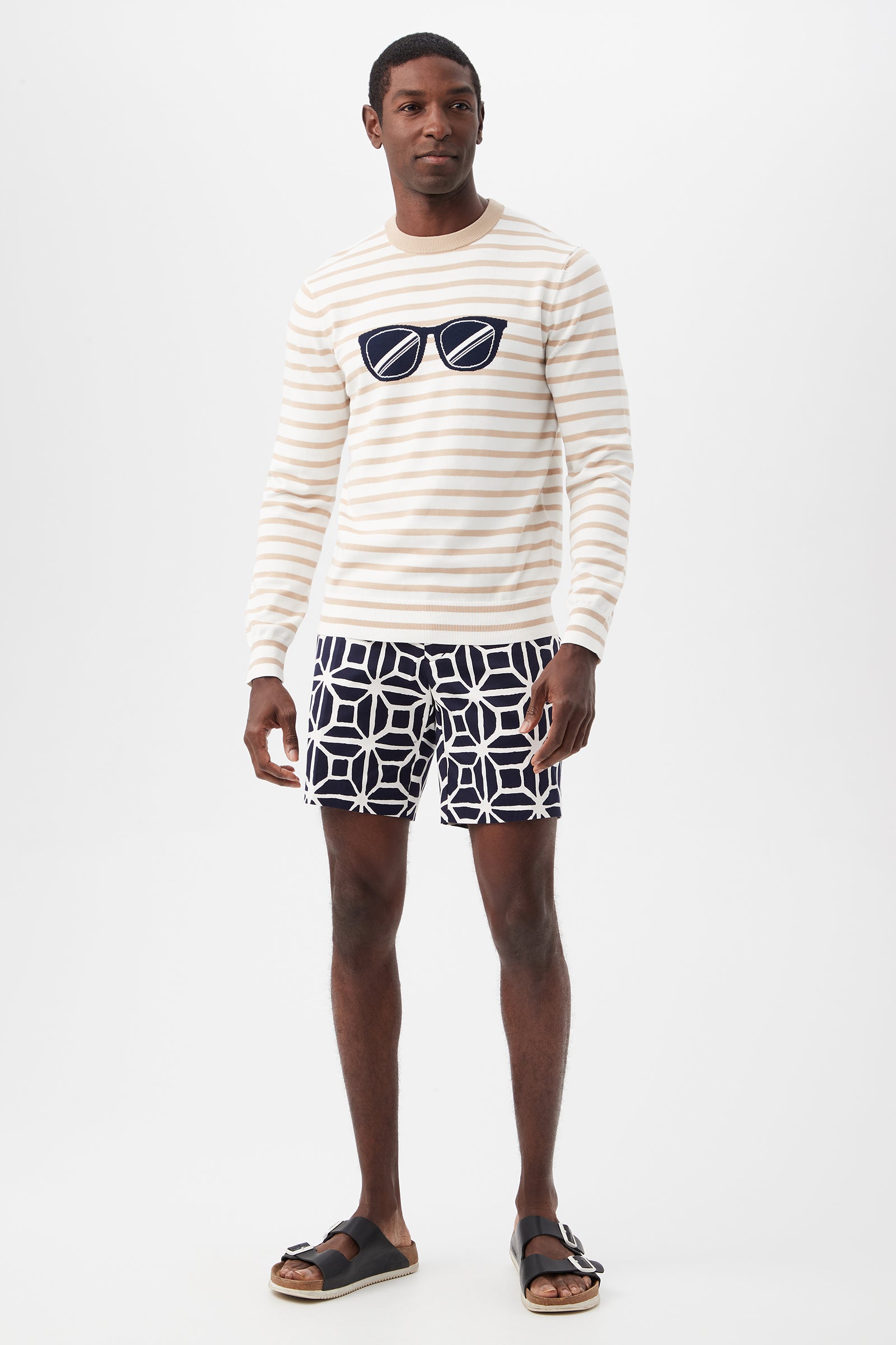 A man stands against a plain background wearing the Julian Sunglasses Crewneck Sweater, featuring cream and beige stripes with a sunglasses graphic, paired with navy patterned shorts and black sandals.